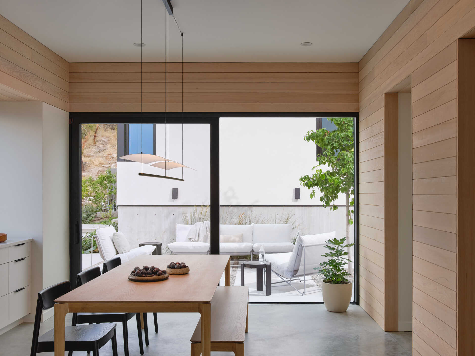 A modern dining room with a large wood table, black chairs, and warm wood-paneled walls opens directly onto a private courtyard. Full-height sliding glass doors connect the dining area to an outdoor lounge with white seating.