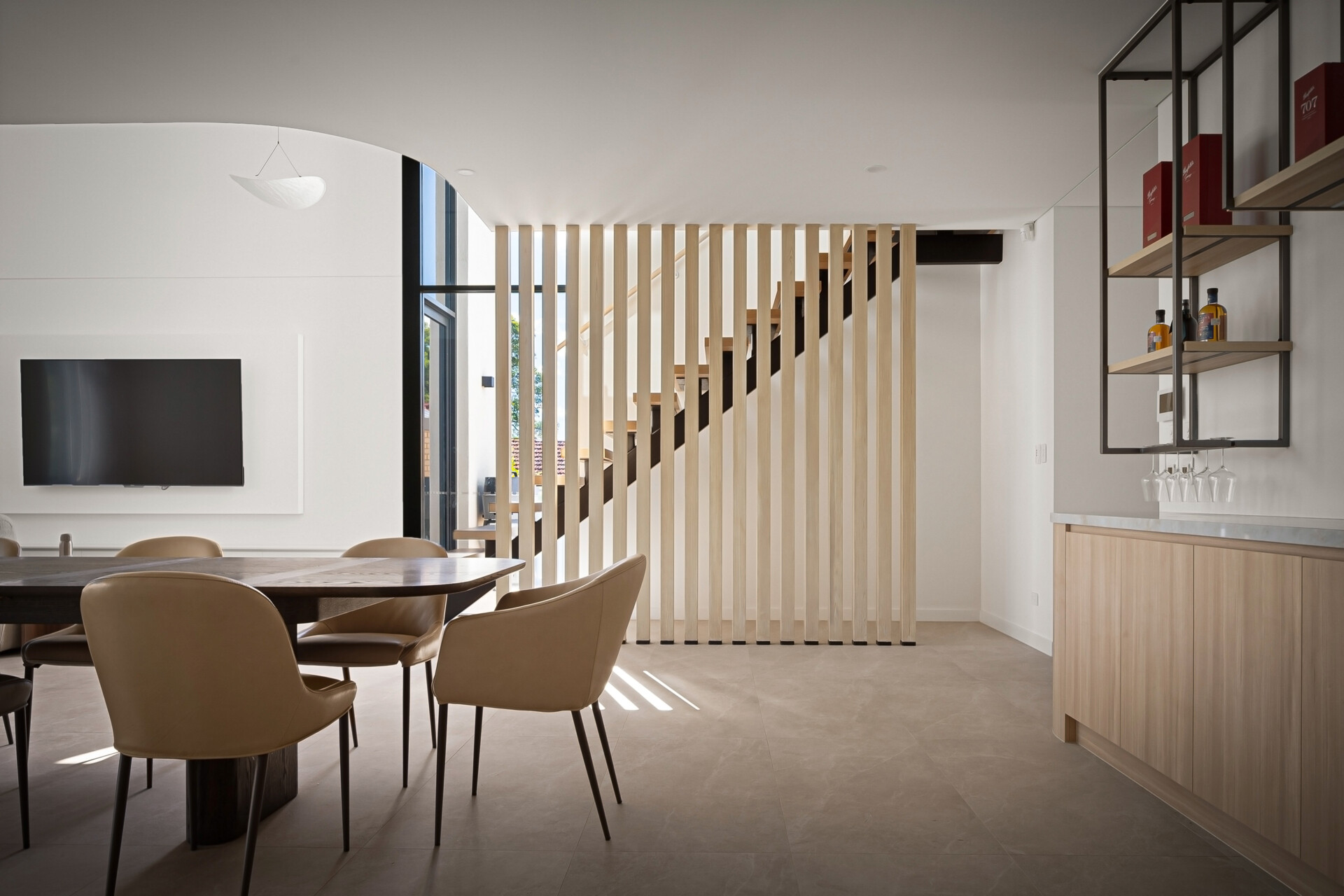 A contemporary dining area with a vertical wood slat partition partially screening an open staircase. A dark dining table, upholstered chairs, and black metal display shelving contrast with the pale flooring and white walls.
