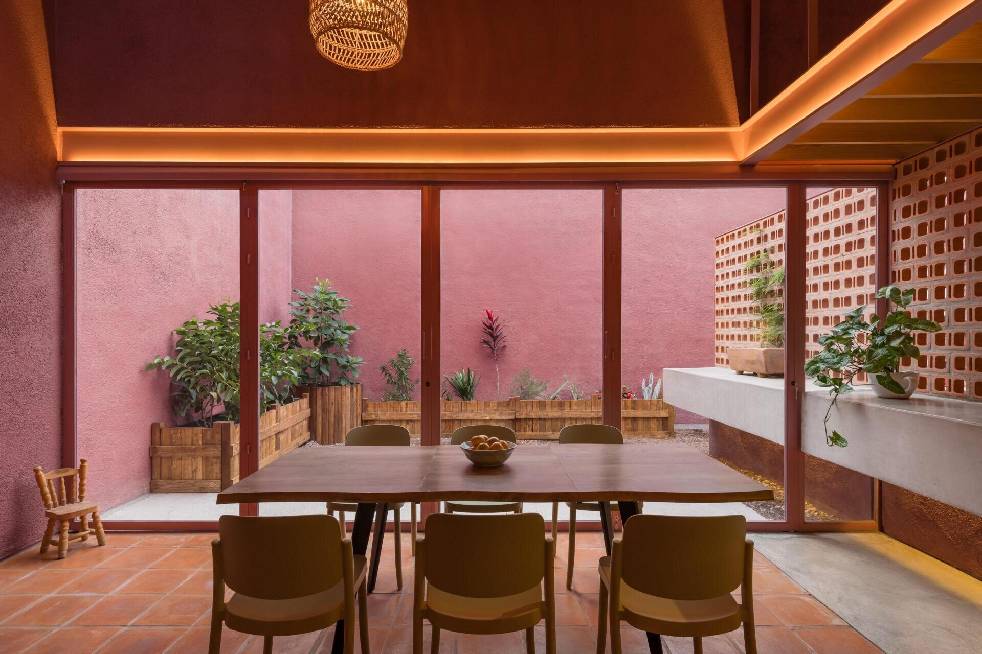 A modern dining room with pink-framed folding glass doors opens to a private courtyard with perforated brick screens, raised garden beds, and pink exterior walls.