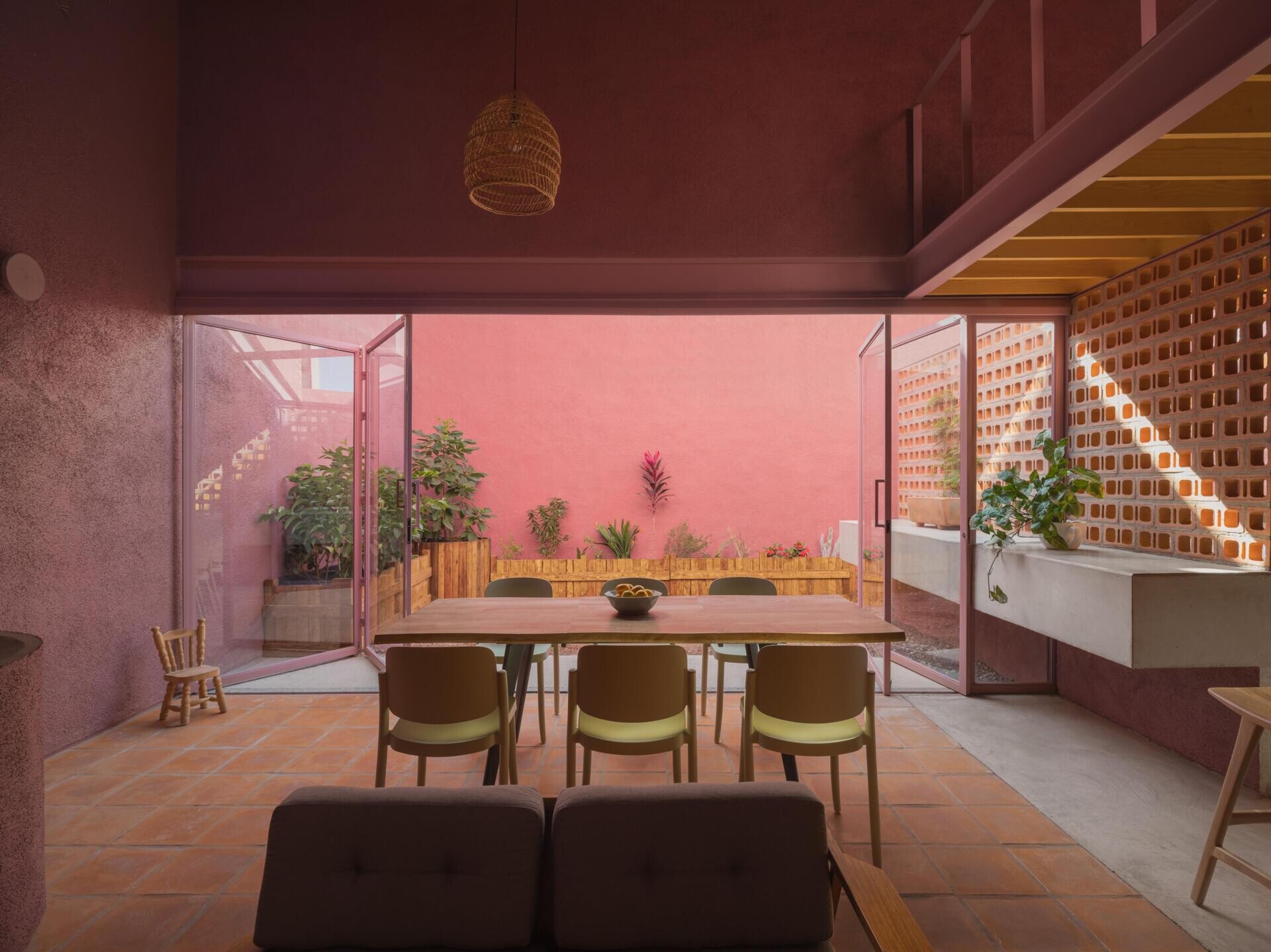 A modern dining room with pink-framed folding glass doors opens to a private courtyard with perforated brick screens, raised garden beds, and pink exterior walls.