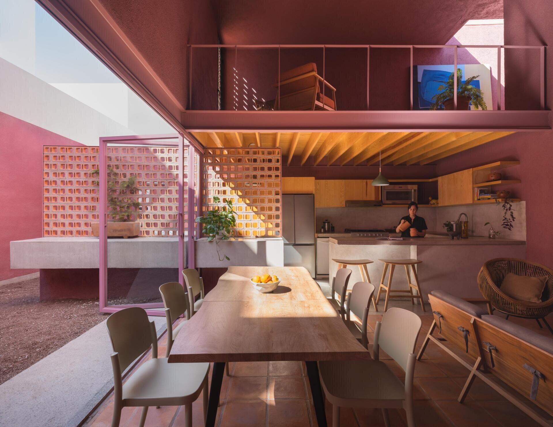 A modern dining room with pink-framed folding glass doors opens to a private courtyard with perforated brick screens, raised garden beds, and pink exterior walls.