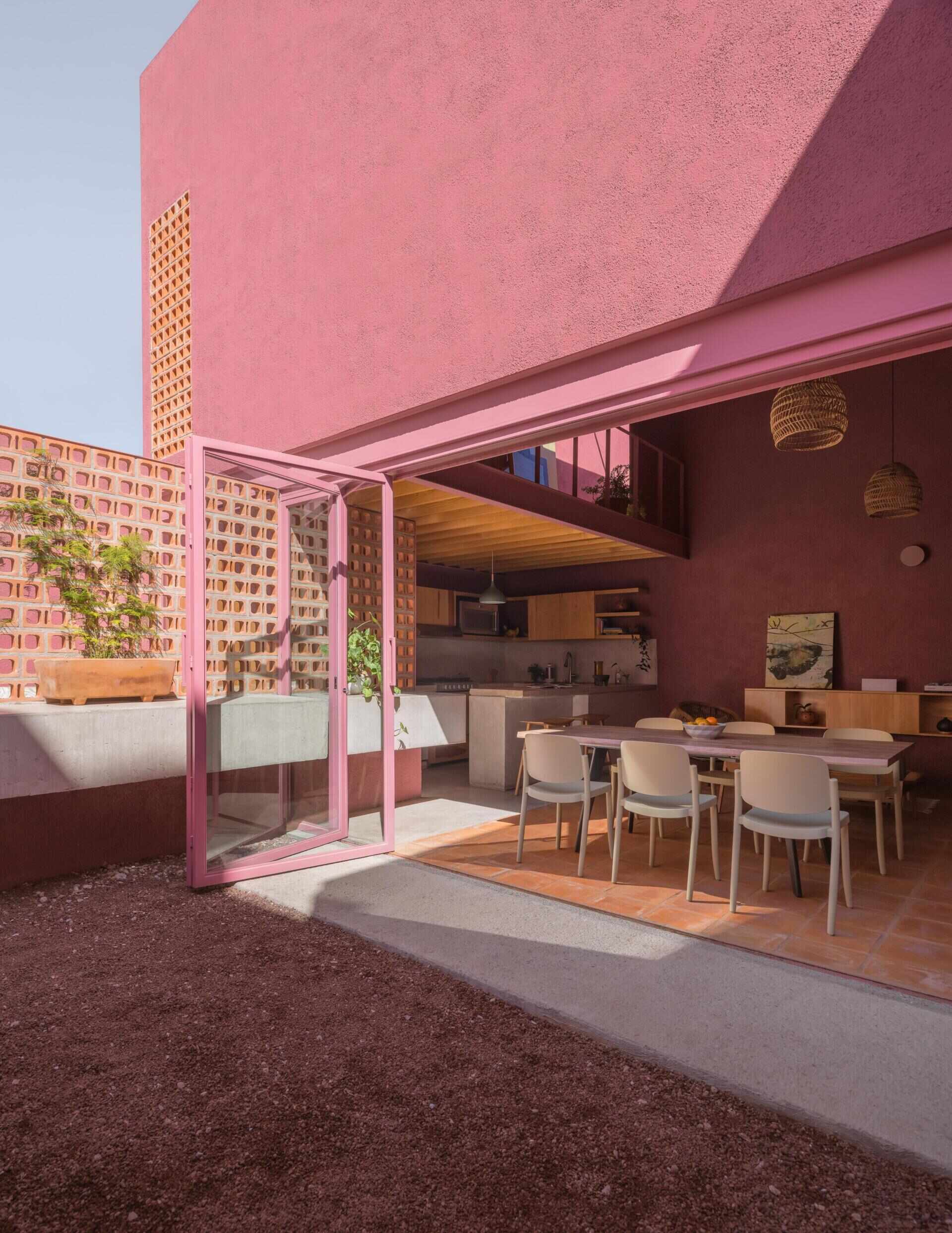 A modern dining room with pink-framed folding glass doors opens to a private courtyard with perforated brick screens, raised garden beds, and pink exterior walls.