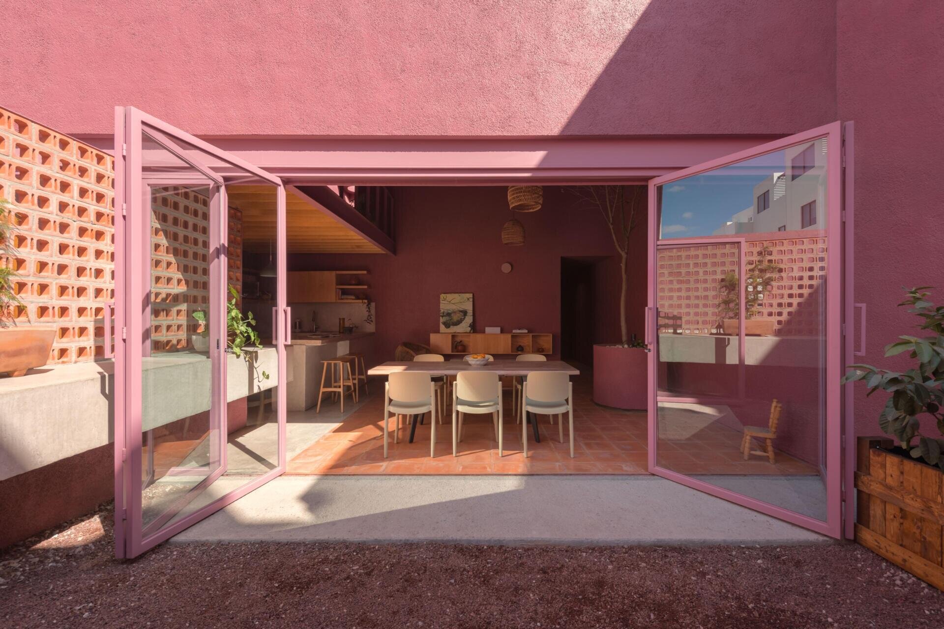 A modern dining room with pink-framed folding glass doors opens to a private courtyard with perforated brick screens, raised garden beds, and pink exterior walls.
