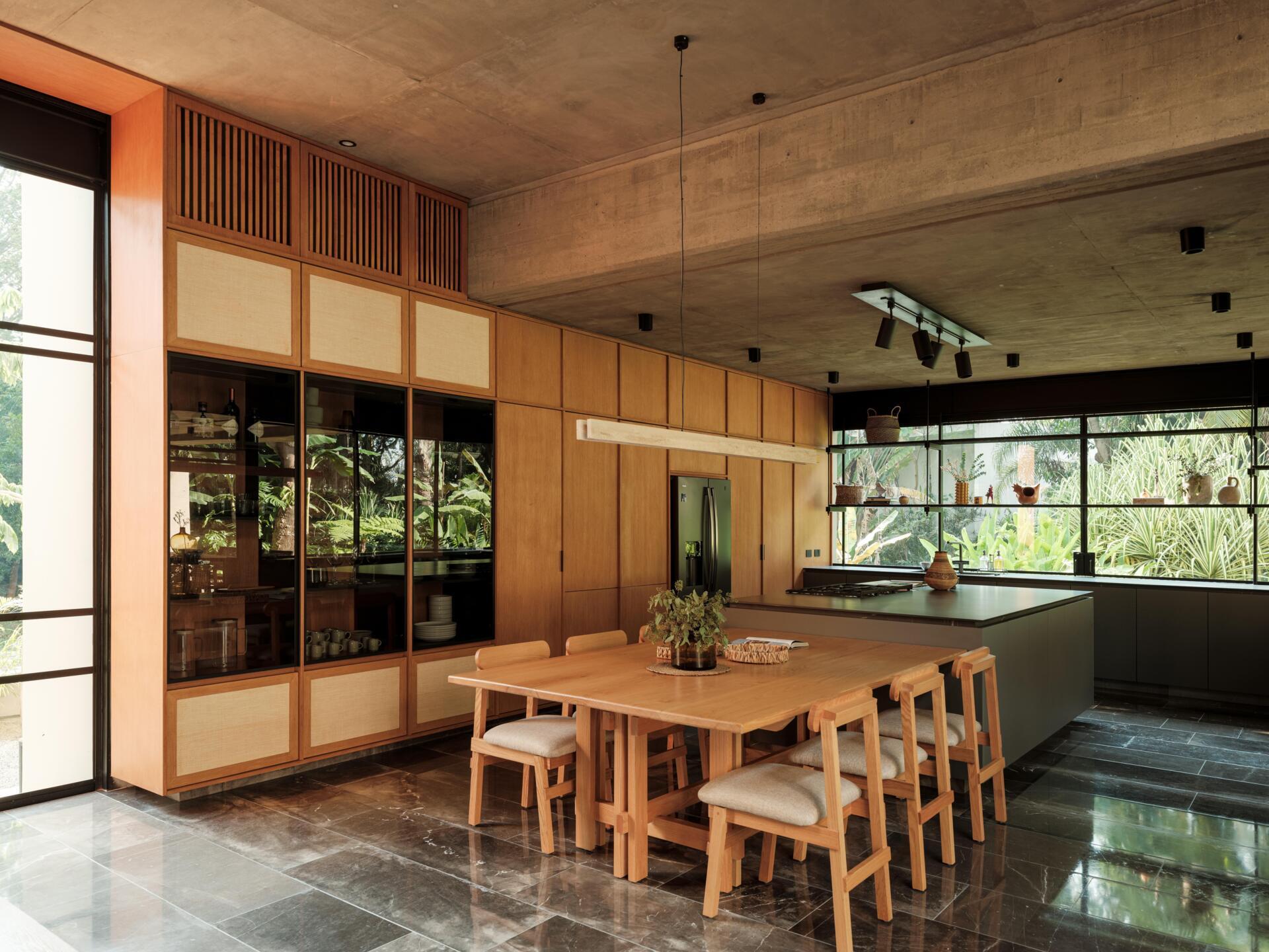 Explore a modern dining room and kitchen in Casa Akina, Mexico, featuring a solid wood dining table, custom timber cabinetry, a dark stone kitchen island, minimalist lighting, and large windows overlooking tropical greenery.