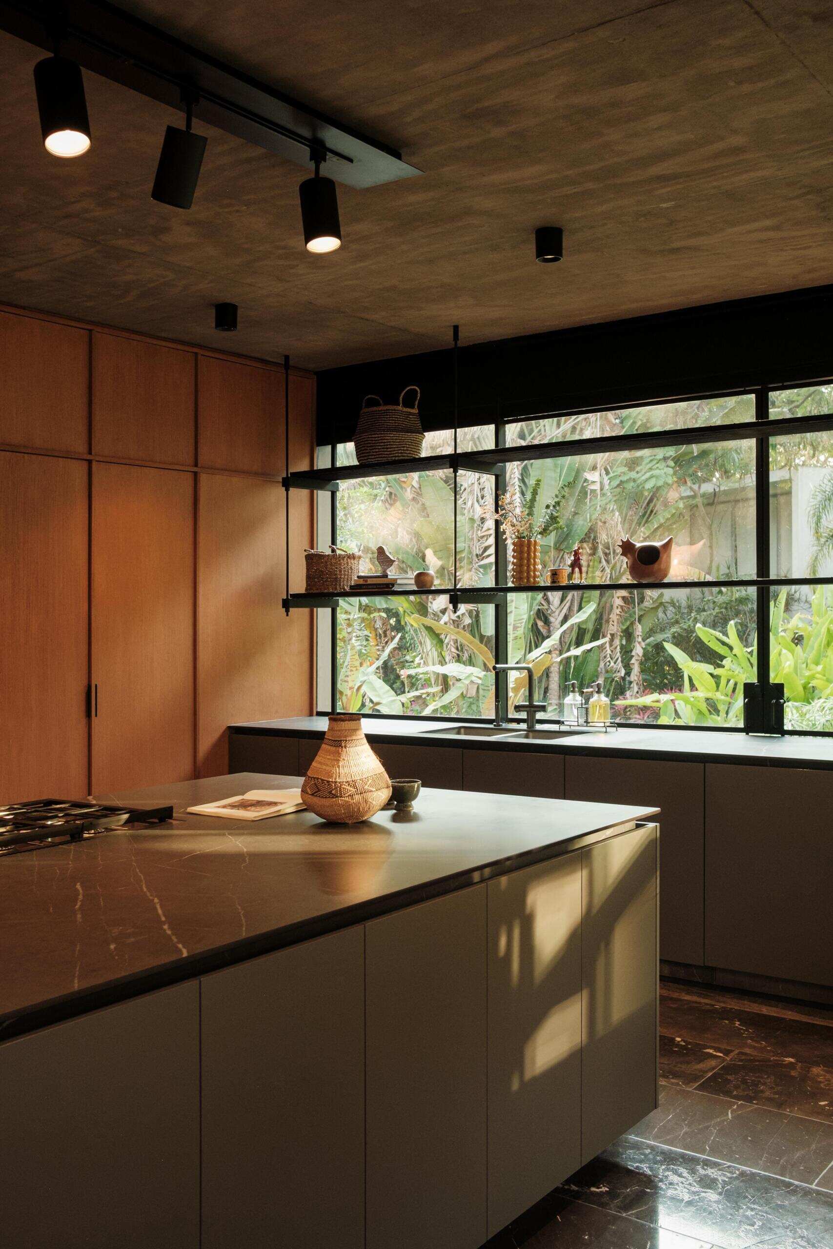 Explore a modern dining room and kitchen in Casa Akina, Mexico, featuring a solid wood dining table, custom timber cabinetry, a dark stone kitchen island, minimalist lighting, and large windows overlooking tropical greenery.
