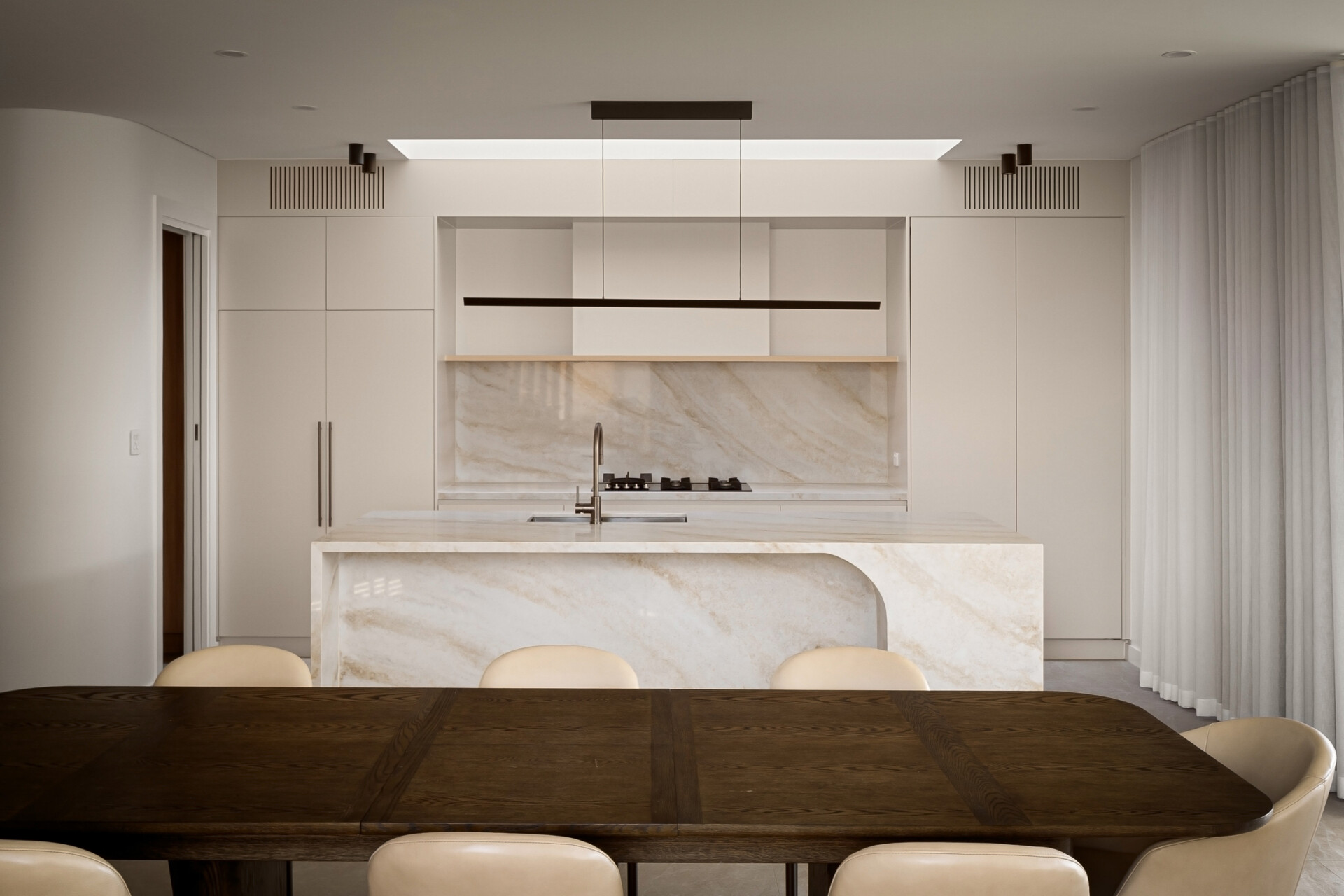 A contemporary kitchen and dining room with a large natural stone island, white floor-to-ceiling cabinets, and a rectangular skylight above the cooking area. A dark wood dining table and cream upholstered chairs create a contrasting dining space.