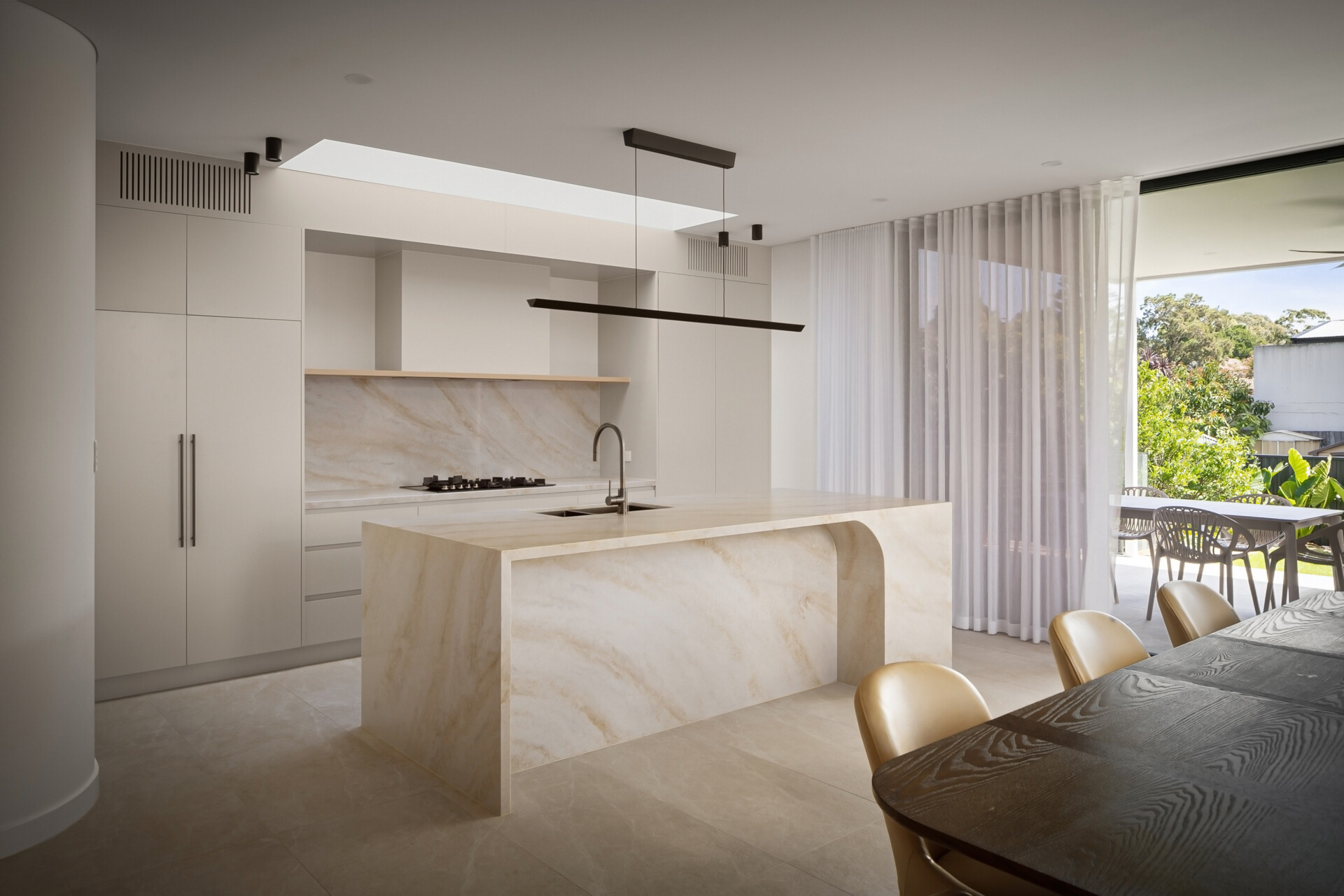 A modern white kitchen with a natural stone island featuring a recessed panel and rounded corner detail. Full-height white cabinetry, a matching stone backsplash, and sheer curtains sit alongside a dark wood dining table, with views toward the outdoor dining area.