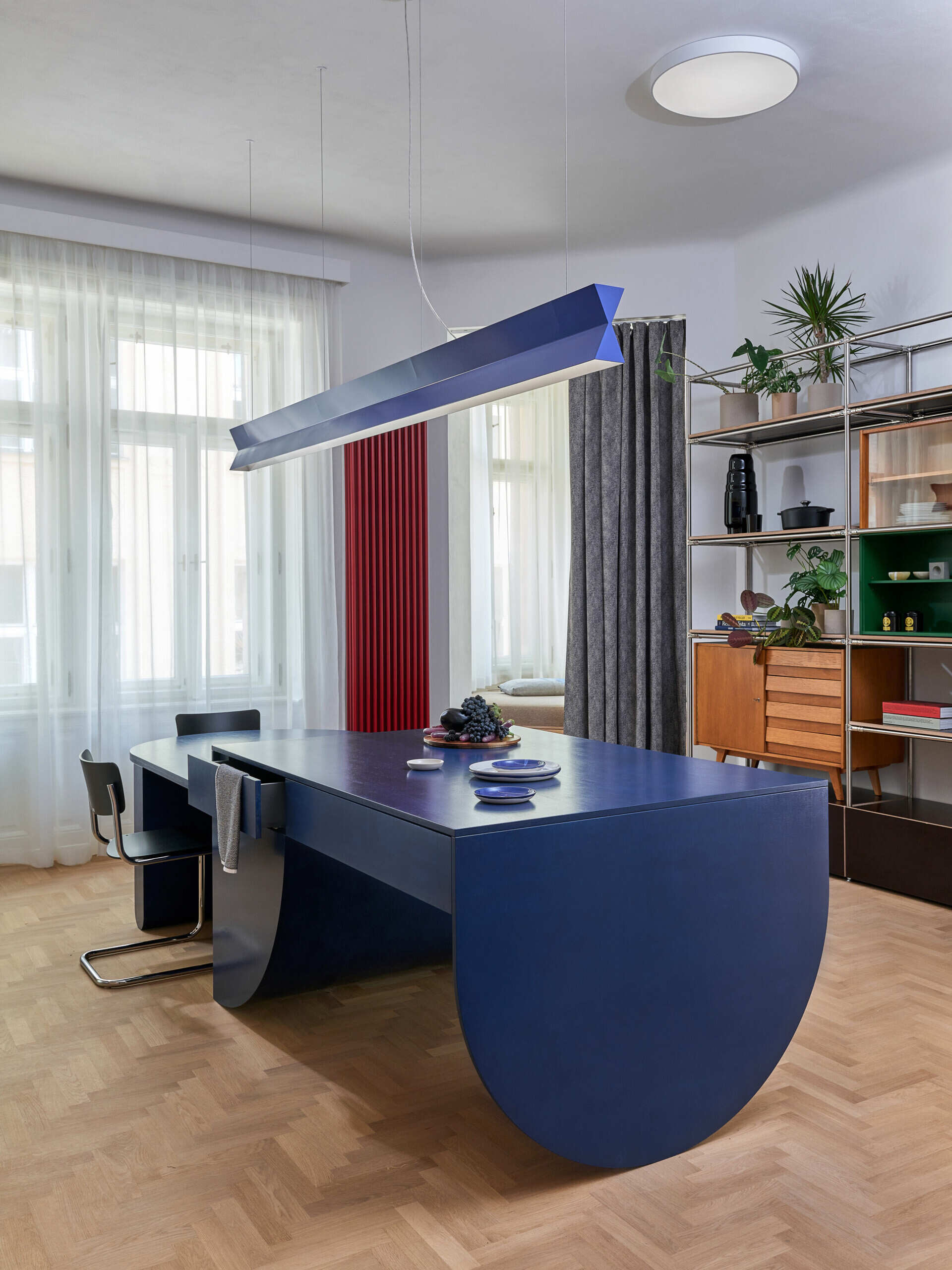 A sculptural blue kitchen island with curved supports creates a distinctive dining and preparation area in a renovated Prague apartment. An angular blue pendant light hangs above, with open metal shelving nearby.