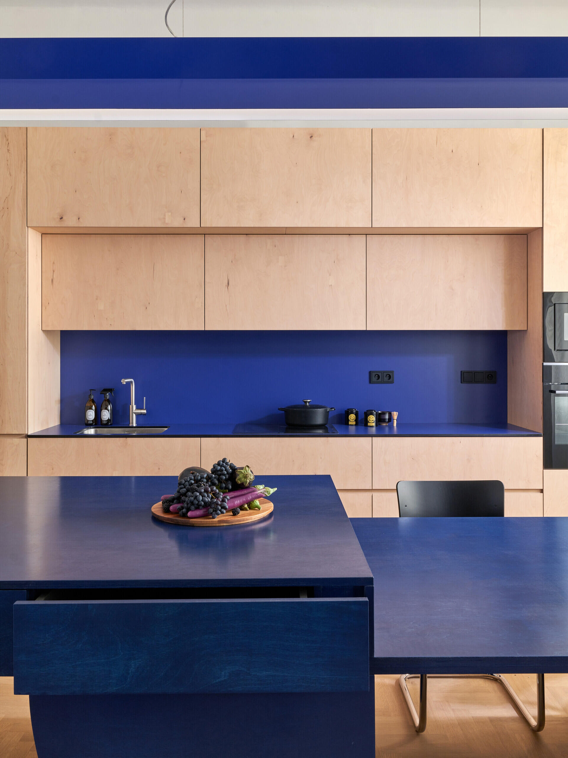 A modern kitchen featuring pale cabinetry, a deep blue backsplash, and a large blue island with an integrated dining surface. The matching blue pendant light adds another geometric element.