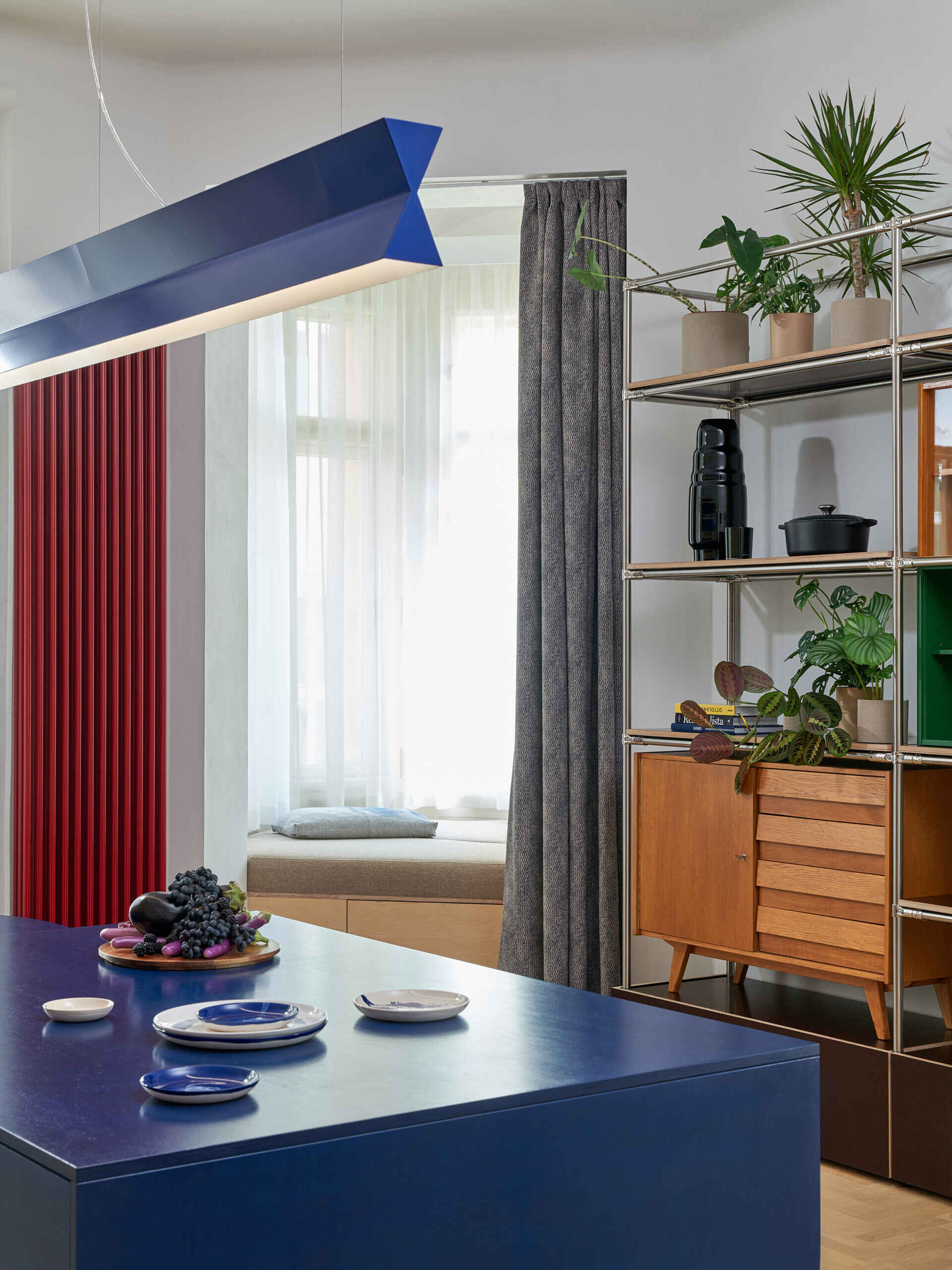 A blue kitchen island sits beneath a long angular pendant light, with a red radiator and floor-to-ceiling curtains nearby. Open metal shelving incorporates vintage-style cabinets and green display compartments.