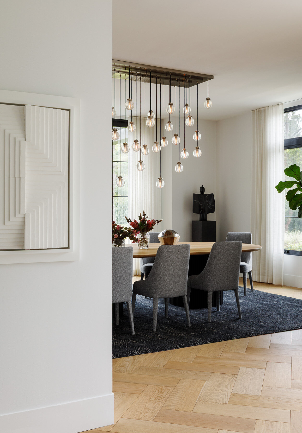 Explore the Atherton Residence formal dining room by Paulina Carcach Studio, featuring a custom two-tone dining table, grey upholstered chairs, cascading glass pendant lights and a collection of large-scale art and sculpture.