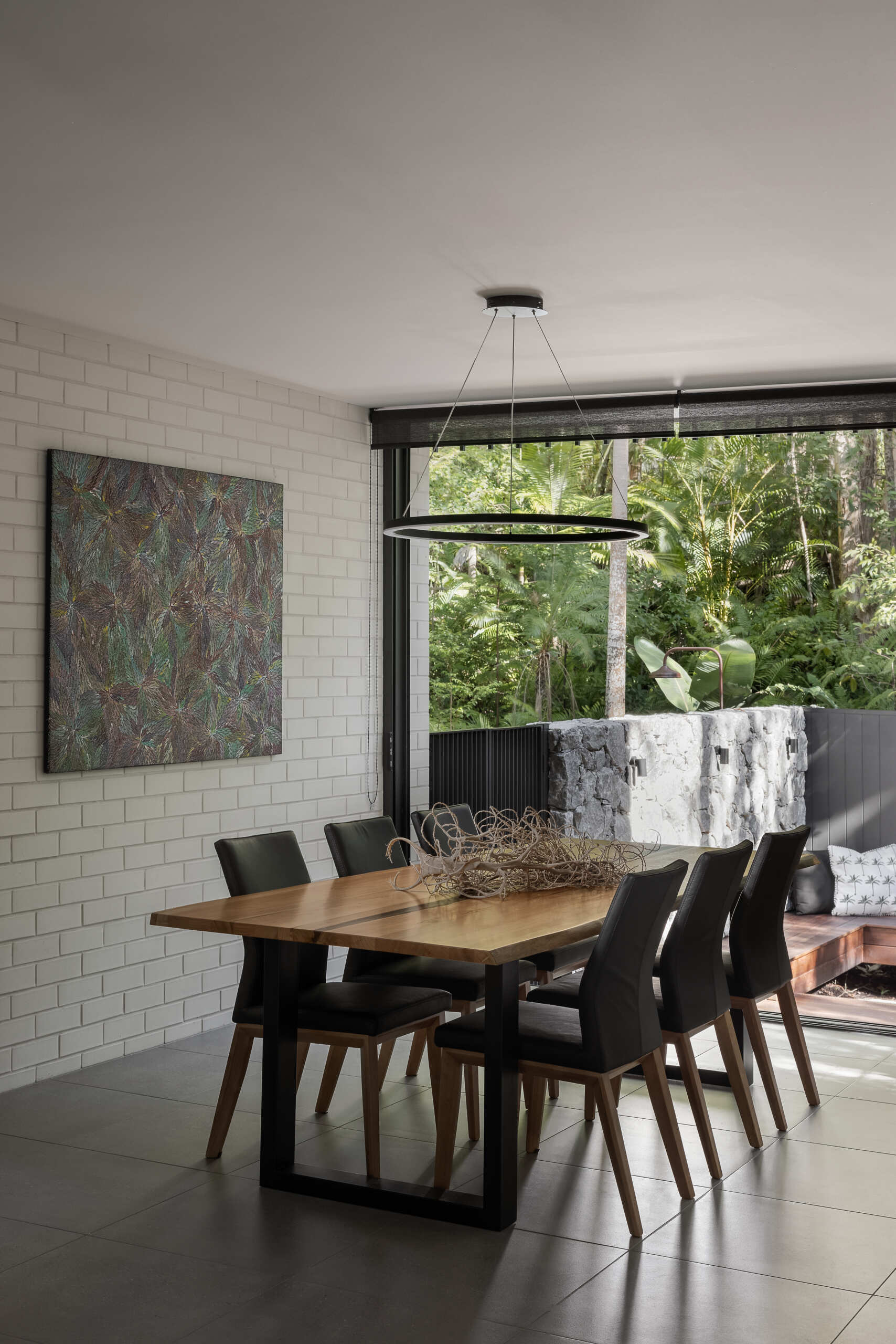 Discover the dining area of Raintree in Noosa Heads, Australia, featuring a natural timber dining table, black upholstered chairs, a modern circular pendant light, botanical artwork, and a built-in window seat overlooking tropical greenery.