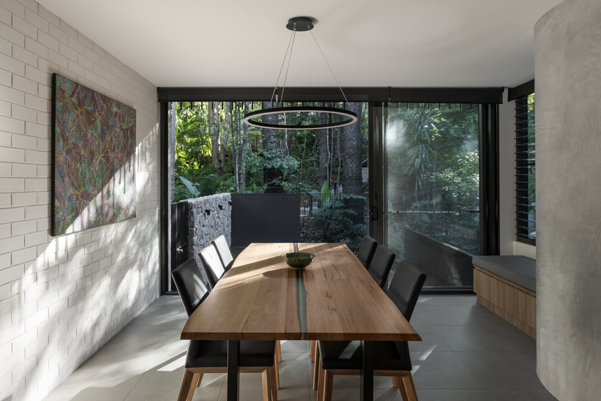 Discover the dining area of Raintree in Noosa Heads, Australia, featuring a natural timber dining table, black upholstered chairs, a modern circular pendant light, botanical artwork, and a built-in window seat overlooking tropical greenery.