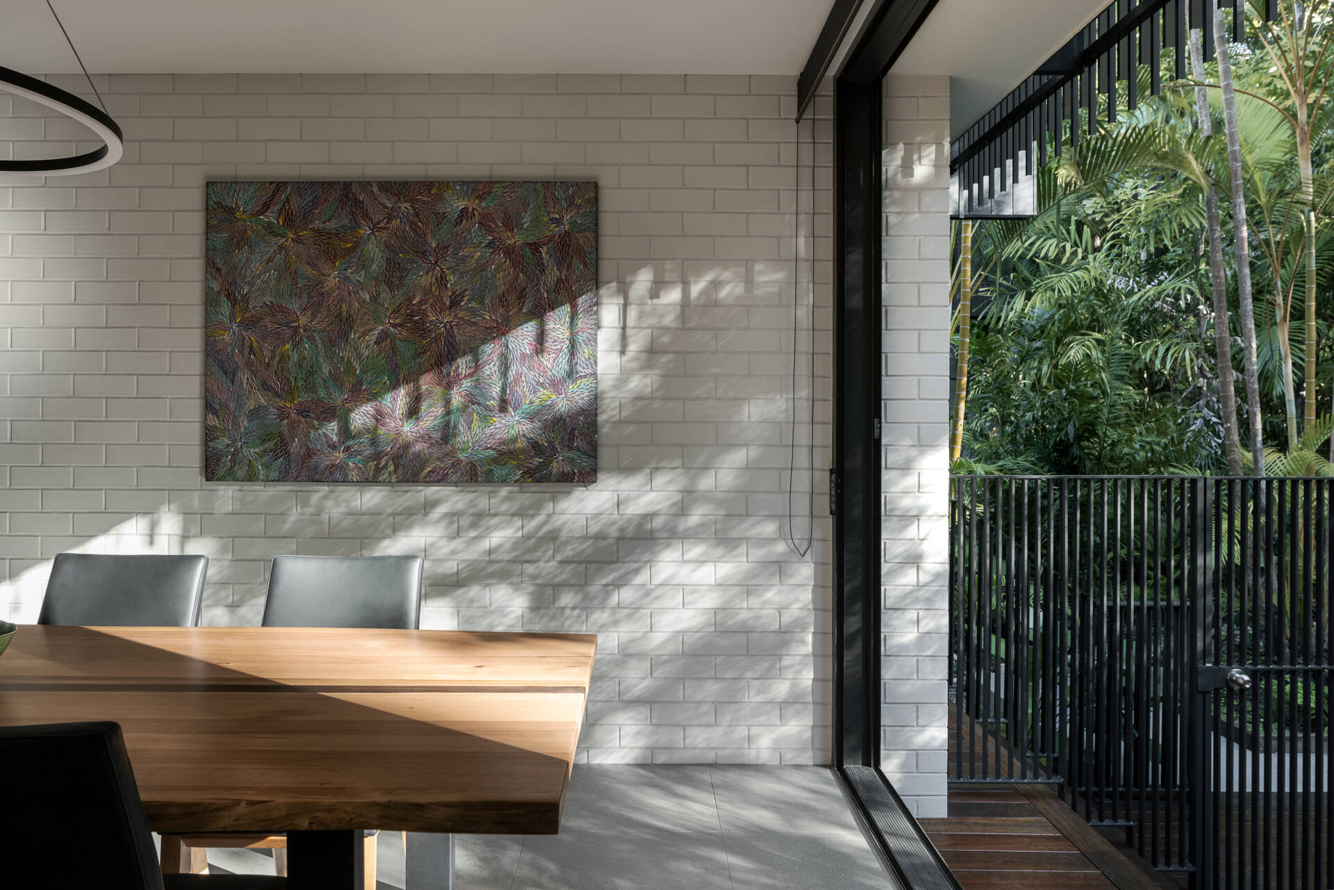 Discover the dining area of Raintree in Noosa Heads, Australia, featuring a natural timber dining table, black upholstered chairs, a modern circular pendant light, botanical artwork, and a built-in window seat overlooking tropical greenery.