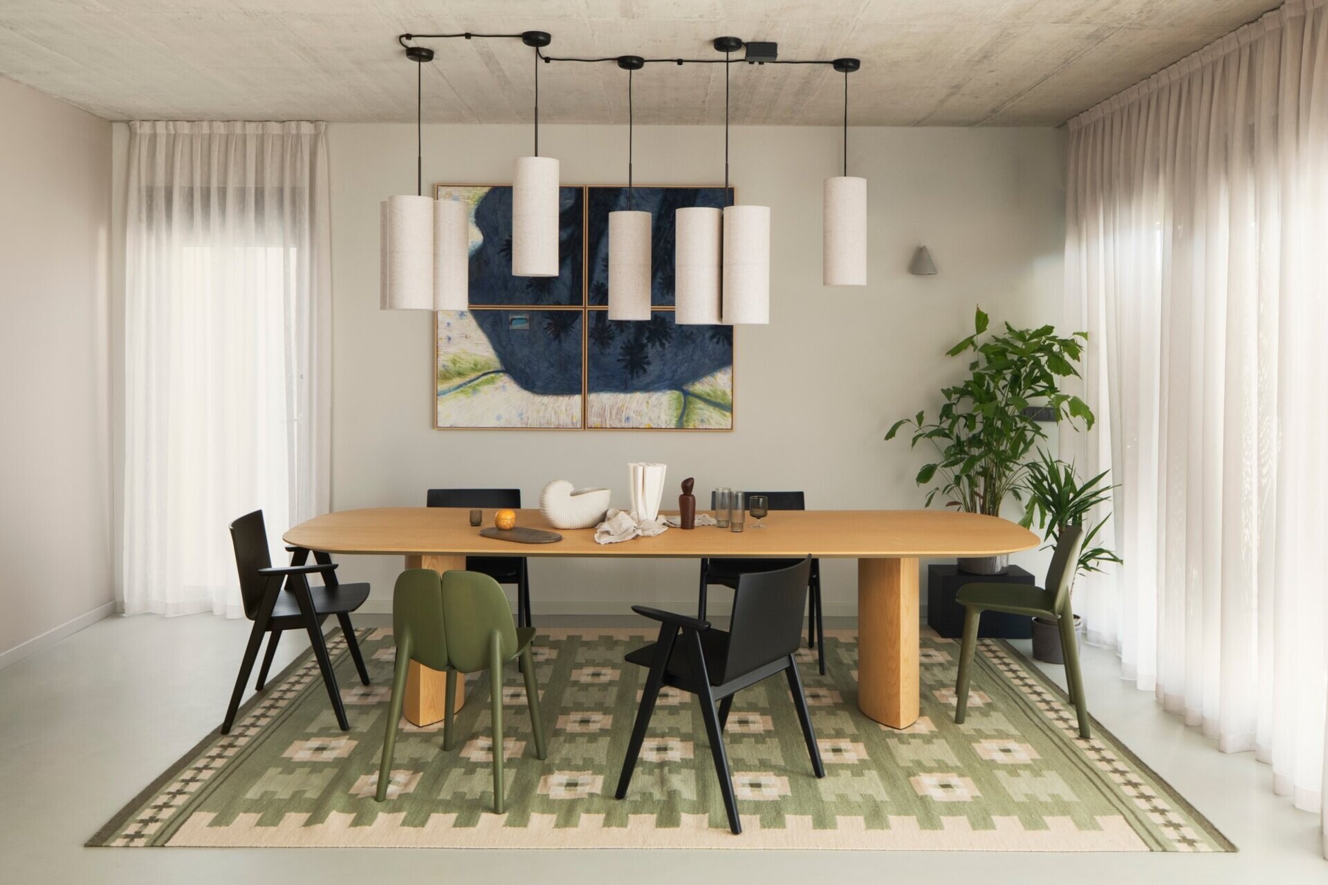 Discover the dining room at Arc House in Croatia, with a long oak-toned dining table, mismatched green and black chairs, patterned rug, sculptural pendant lighting and colourful contemporary artwork.