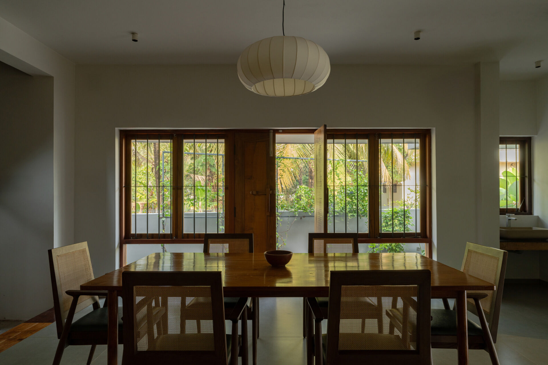 A modern dining room in Kerala features a large wooden table, six cane-back dining chairs, a white pendant light, and garden-facing windows.