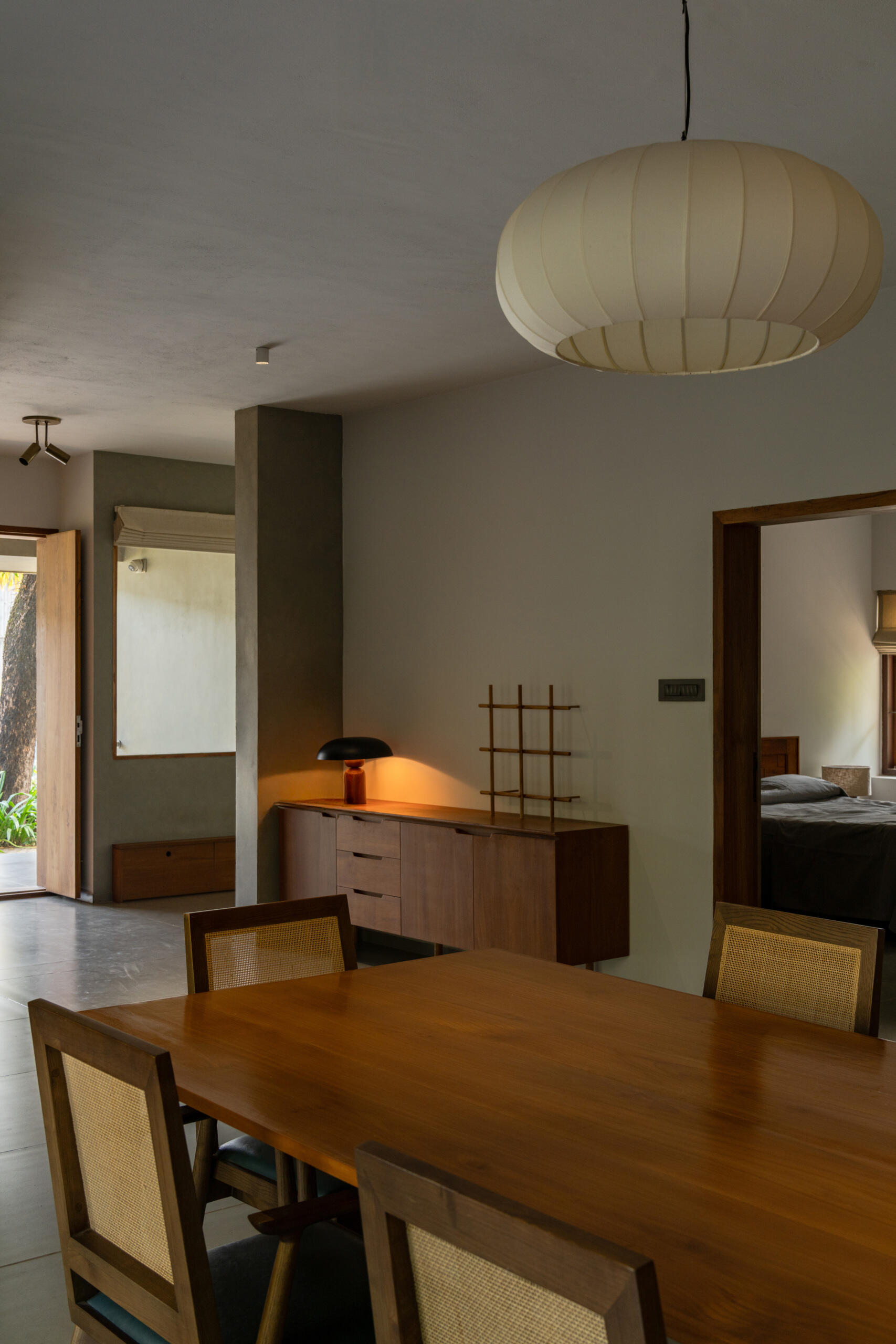 A modern dining room in Kerala features a large wooden table, six cane-back dining chairs, a white pendant light, and garden-facing windows.