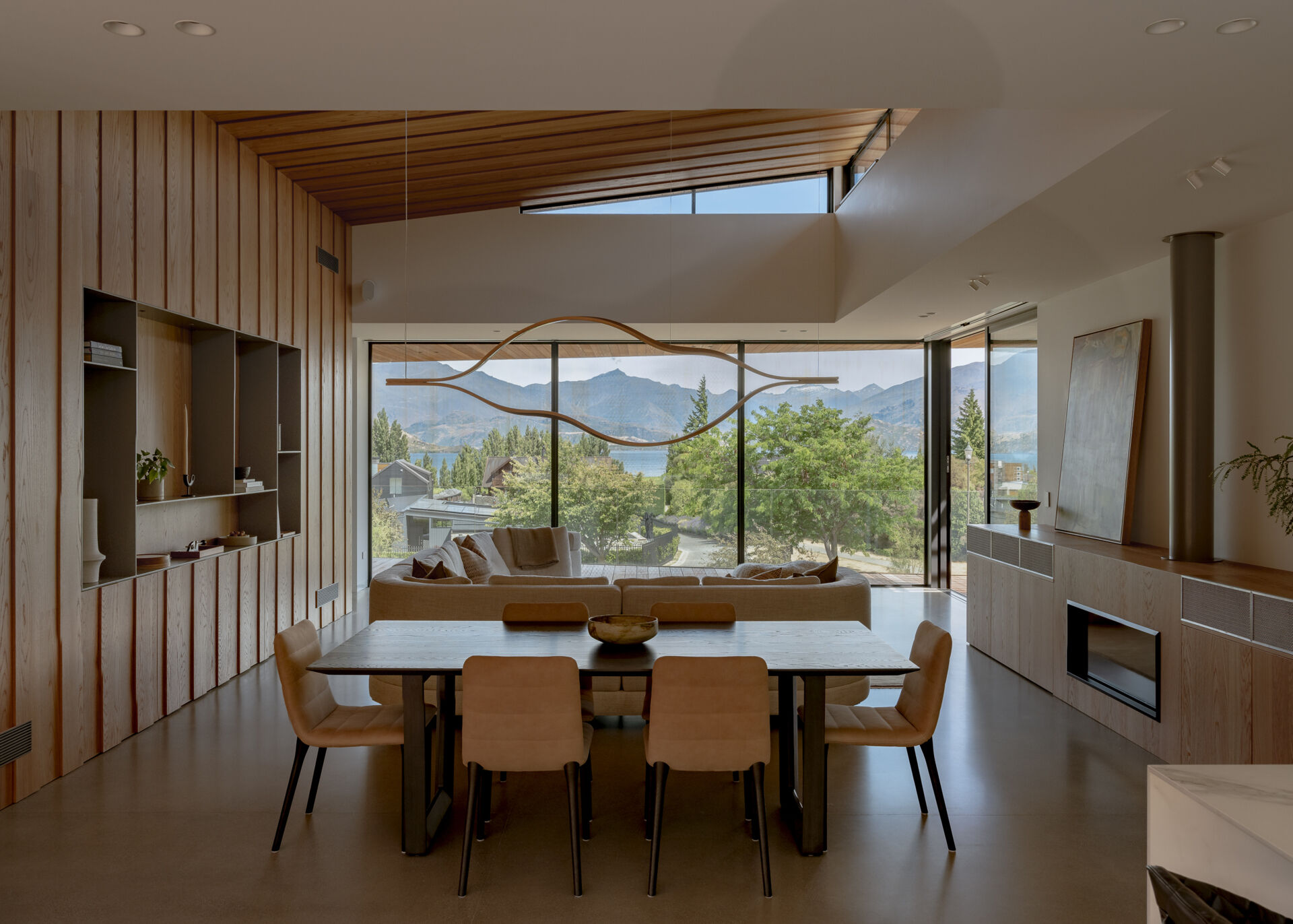 Contemporary dining room with a rectangular dining table, upholstered chairs, sculptural pendant light, and floor-to-ceiling windows overlooking Lake W?naka. Warm wood wall paneling and an angled ceiling add texture to the open-plan interior.
