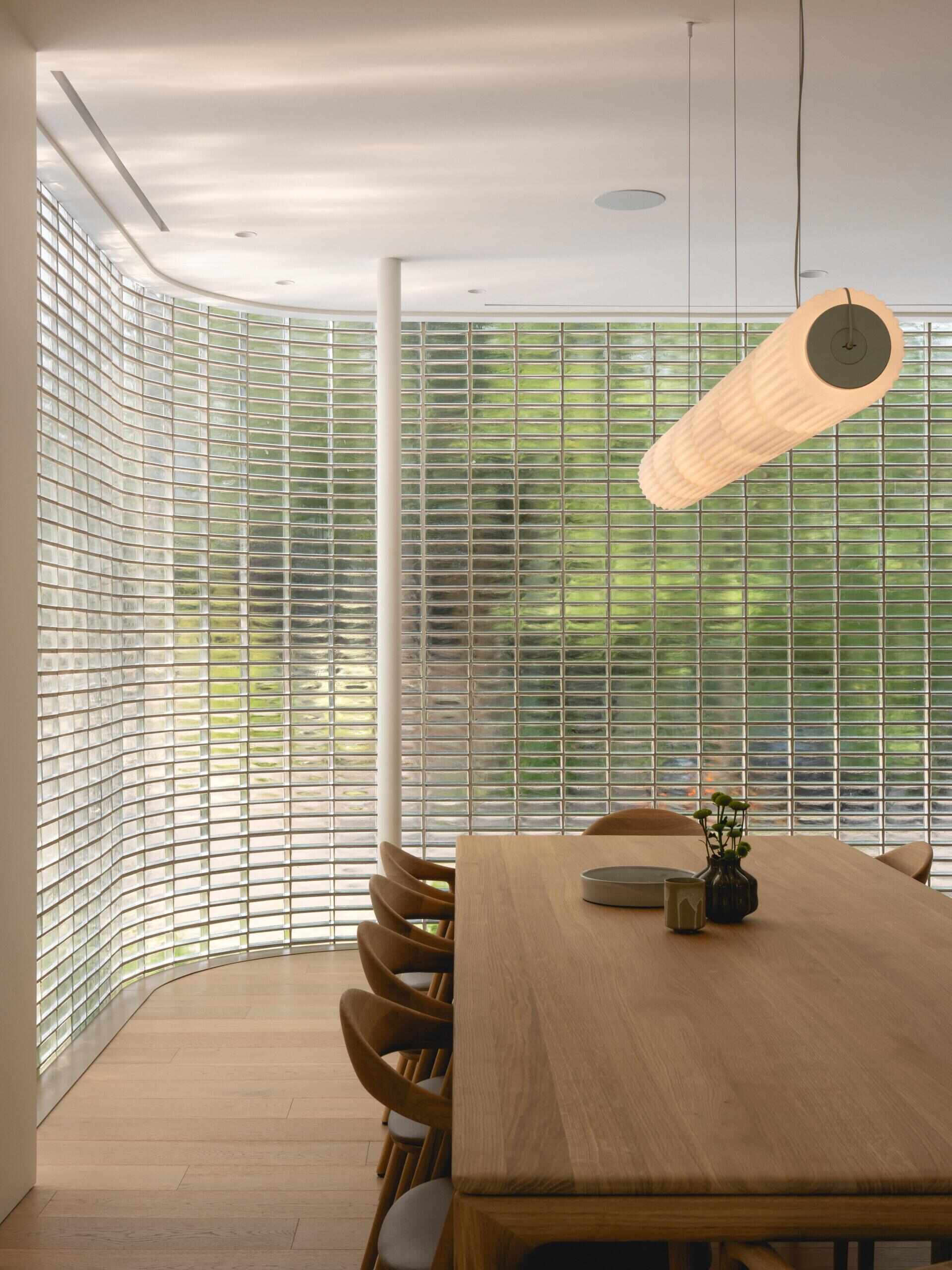Discover the curved glass-brick dining room at The Lens, a contemporary Montreal home by MU Architecture. A warm timber dining table, sculptural pendant lighting, and translucent glass walls create a beautiful minimalist interior.