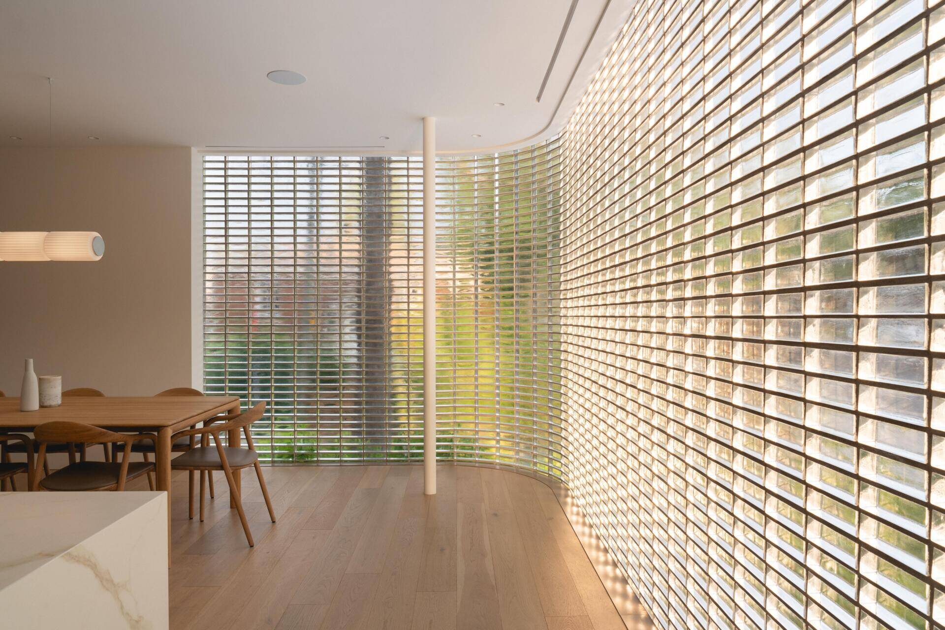 Discover the curved glass-brick dining room at The Lens, a contemporary Montreal home by MU Architecture. A warm timber dining table, sculptural pendant lighting, and translucent glass walls create a beautiful minimalist interior.