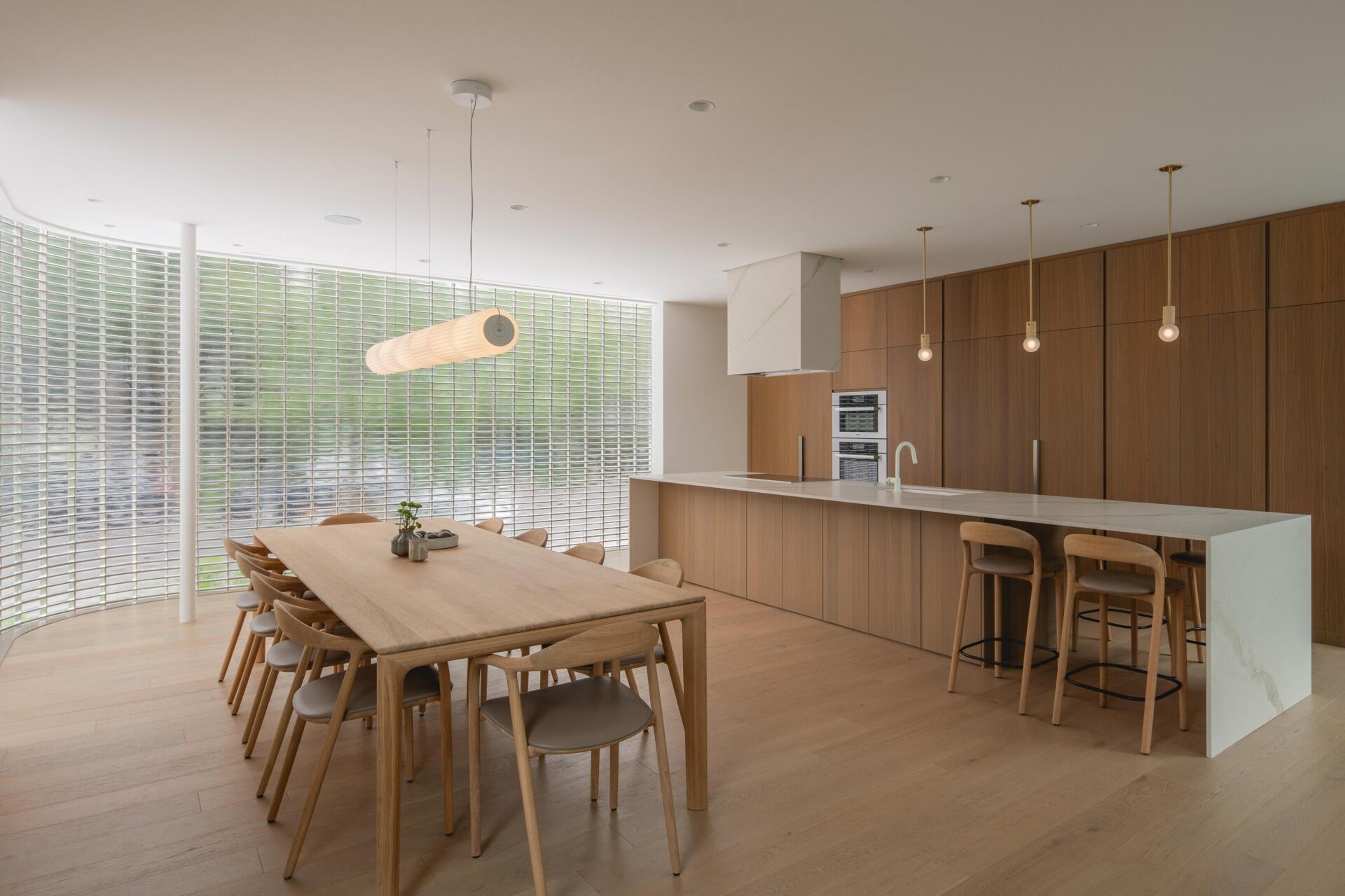 Explore a modern walnut kitchen in The Lens by MU Architecture. Full-height wood cabinetry, a minimalist porcelain island, sculptural range hood, brass pendant lighting, and wooden bar stools define this Montreal home's contemporary design.