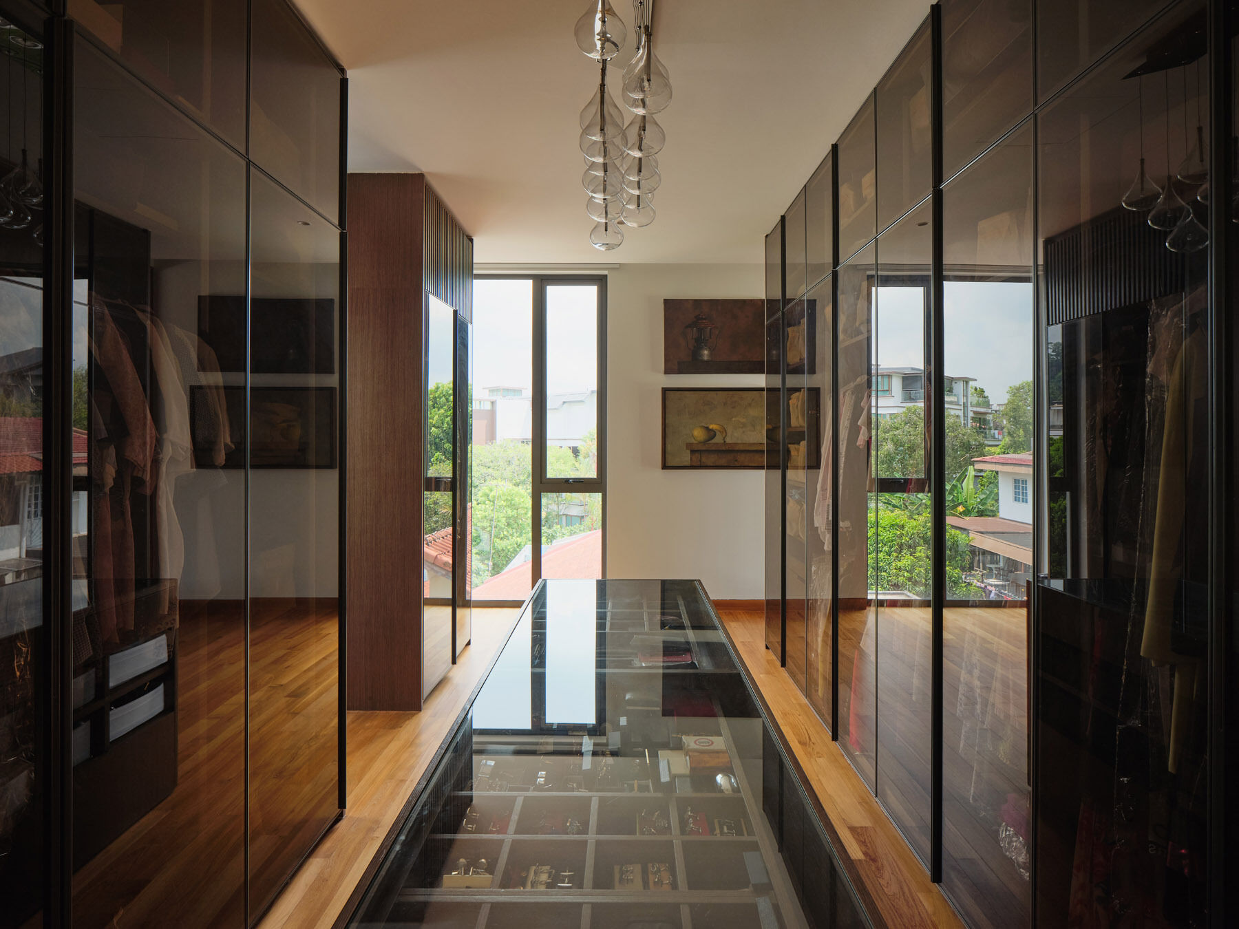 Discover a contemporary walk-in dressing room with floor-to-ceiling smoked-glass wardrobes, a central glass-topped storage island, timber floors, and elegant pendant lighting.