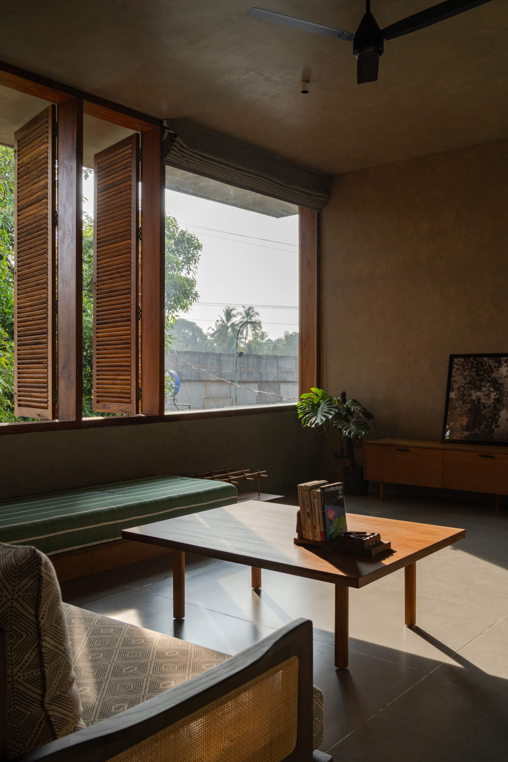 An upper-floor entertainment room with wooden shutters, a green daybed, a built-in desk, and treetop views shows how contemporary design can embrace its natural surroundings.