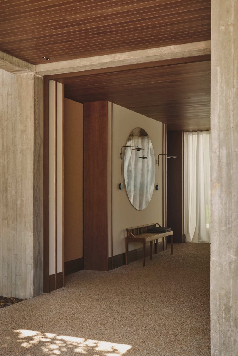 Step inside Casa Tossal and explore its modern entryway, featuring floating-style concrete garden steps, wood ceilings, large wooden doors, sculptural artwork, and a minimalist wooden bench.