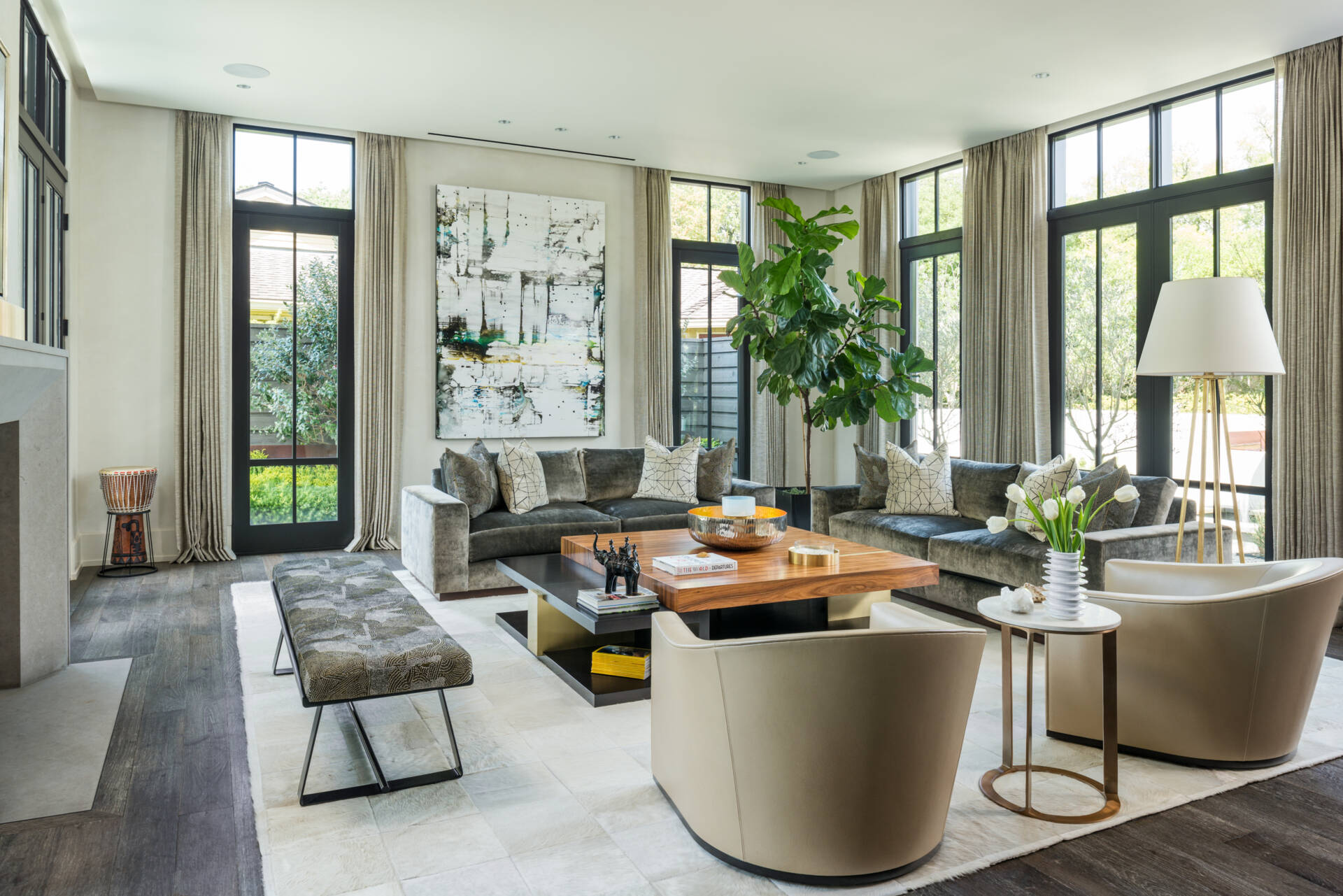 This contemporary living room features gray velvet sofas, curved cream armchairs, tall black-framed windows, abstract artwork, dark wood floors, and a large central coffee table.