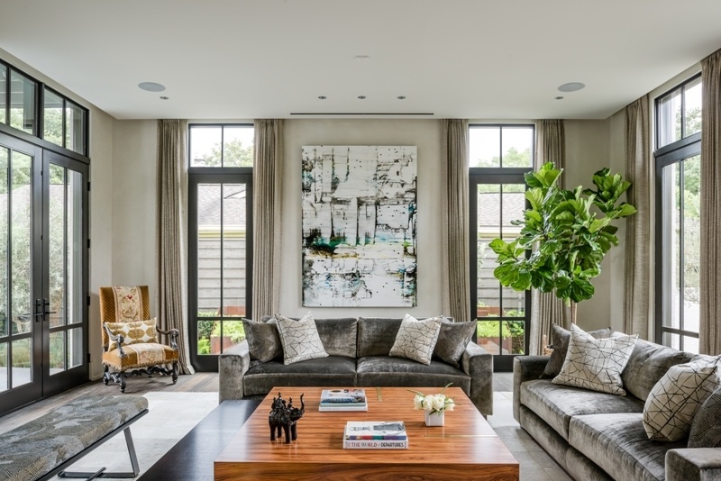 This contemporary living room features gray velvet sofas, curved cream armchairs, tall black-framed windows, abstract artwork, dark wood floors, and a large central coffee table.