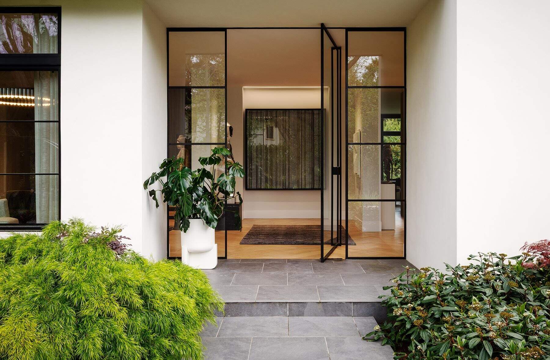Step inside the Atherton Residence through a black-framed glass front door, where grey stone paving, lush planting and a prominent artwork create a clean transition from garden to interior.