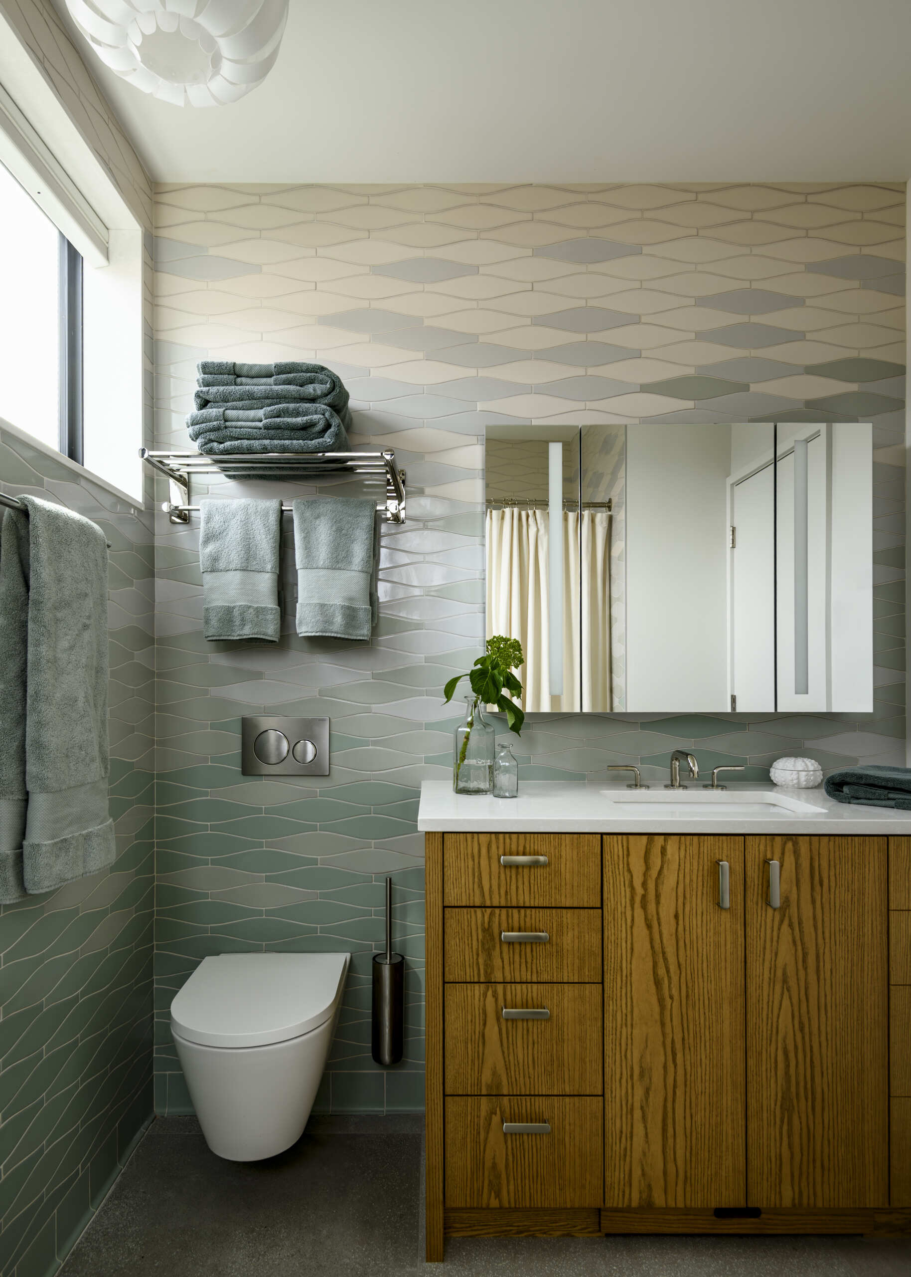 Featuring wave-shaped tiles in shades of white, blue, and seafoam green, a warm wood vanity, wall-mounted toilet, and modern fixtures, this small bathroom makes the most of its compact layout.