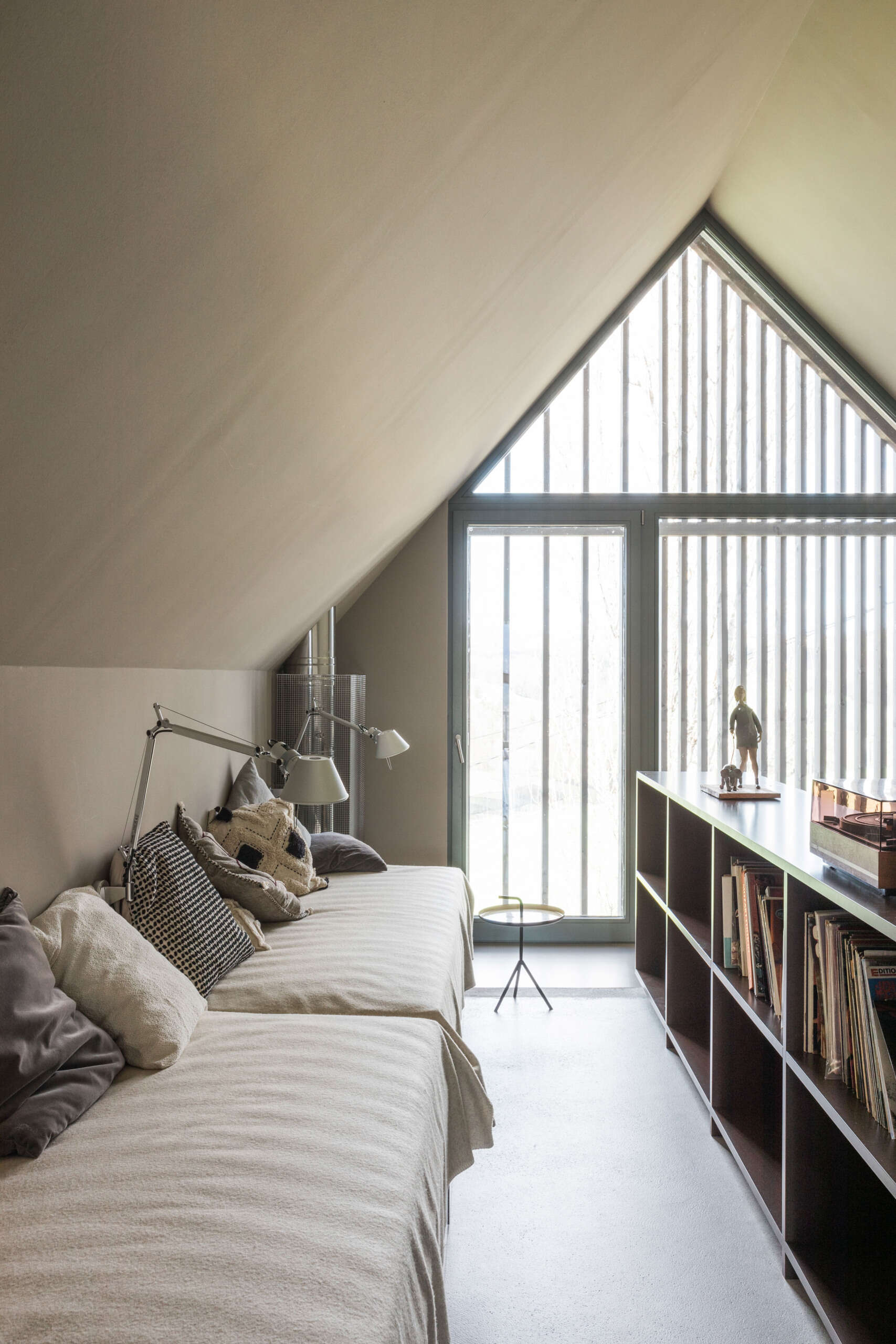 An upper-floor gallery lounge beneath a pitched roof features daybed seating, a record collection, bookshelves, and a large triangular window with timber screening.