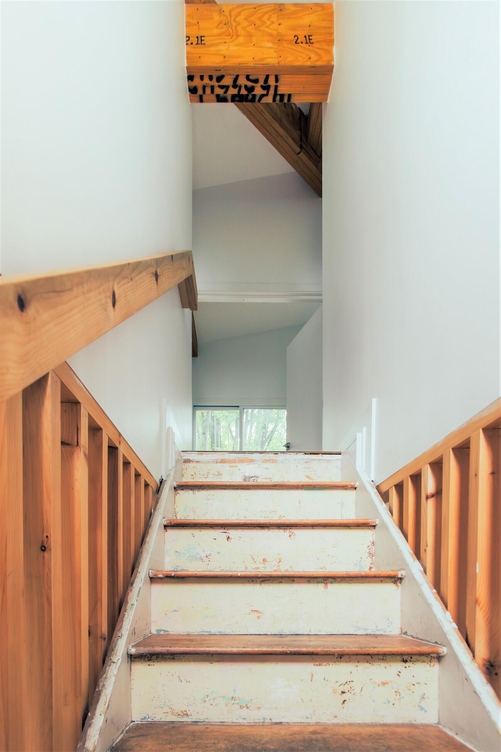 An original wooden staircase becomes an architectural feature in this renovated home, with weathered steps, new timber slat railings, tall white walls, and natural light.