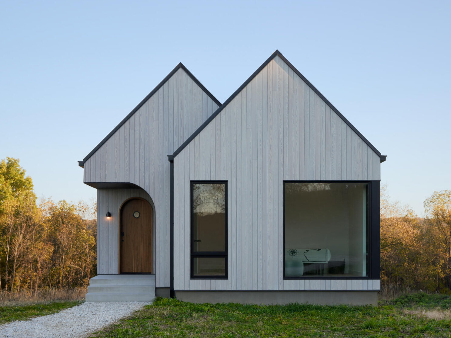 A modern cabin in rural Iowa features twin gabled roofs, vertical wood siding, a recessed arched walnut front door, and black-framed windows overlooking the grassy landscape.