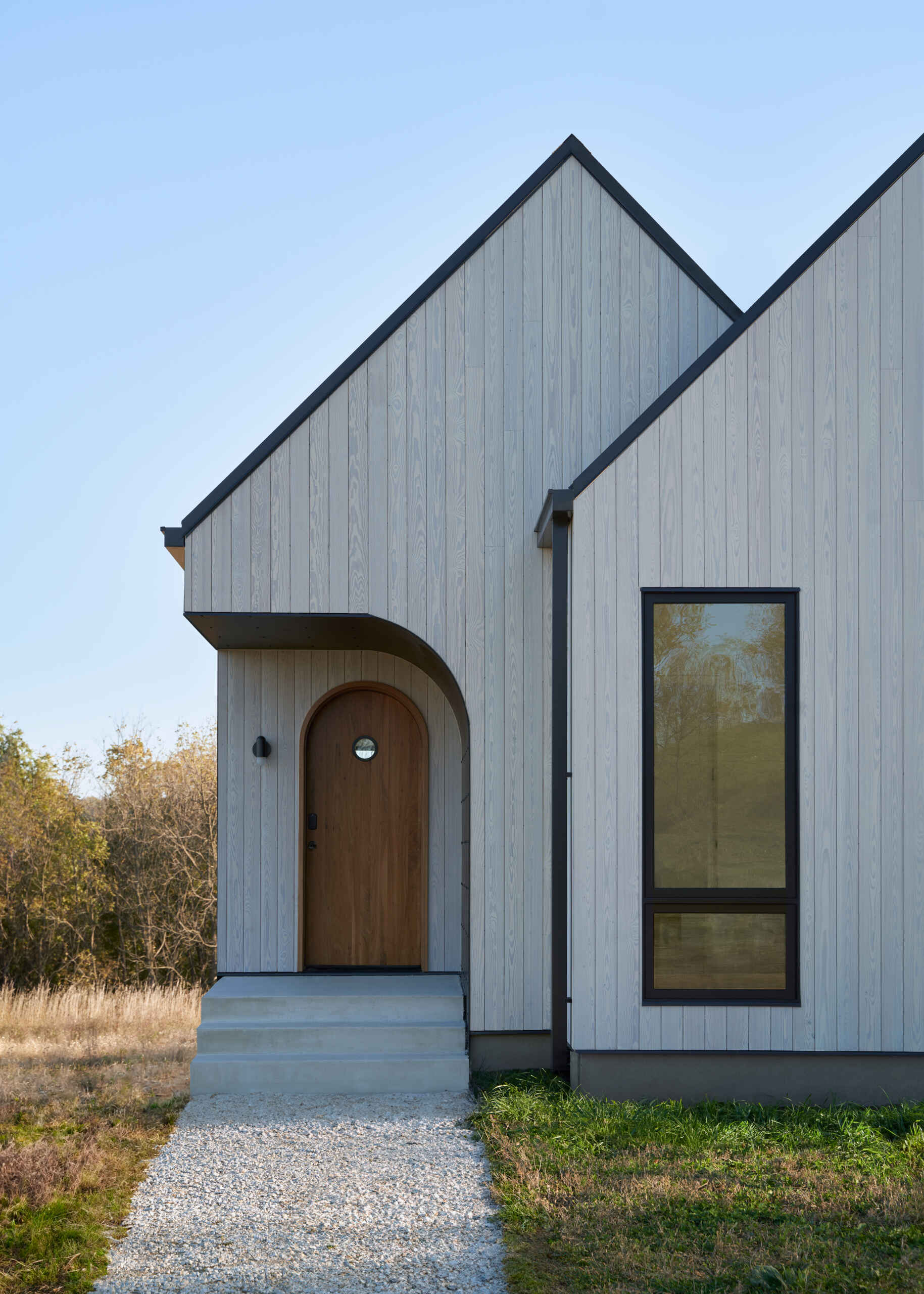 An arched wood front door sits beneath a curved, recessed porch on a modern white house. The sweeping dark metal overhang contrasts with the angular rooflines, vertical siding, and rectangular window.