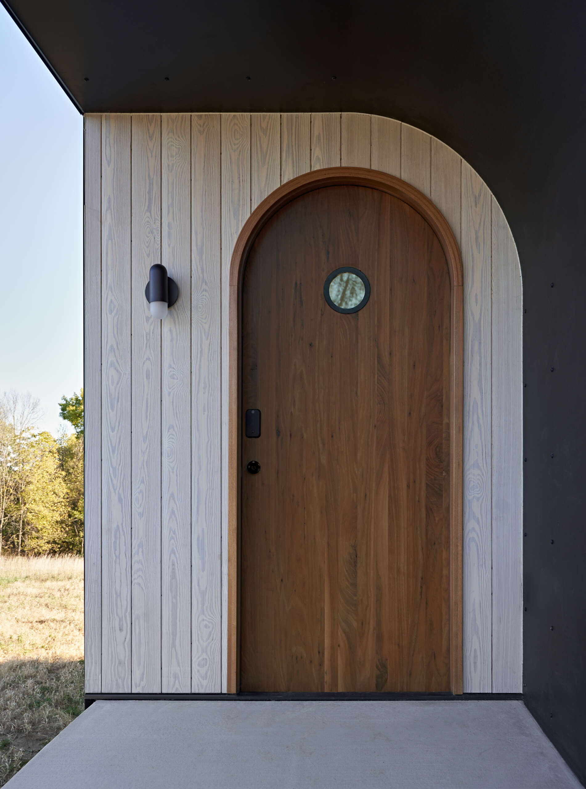 Close-up of an arched walnut front door with a small circular window, curved wood frame, and contrasting pale vertical siding. A black metal-lined recess and modern exterior wall light complete the entrance.