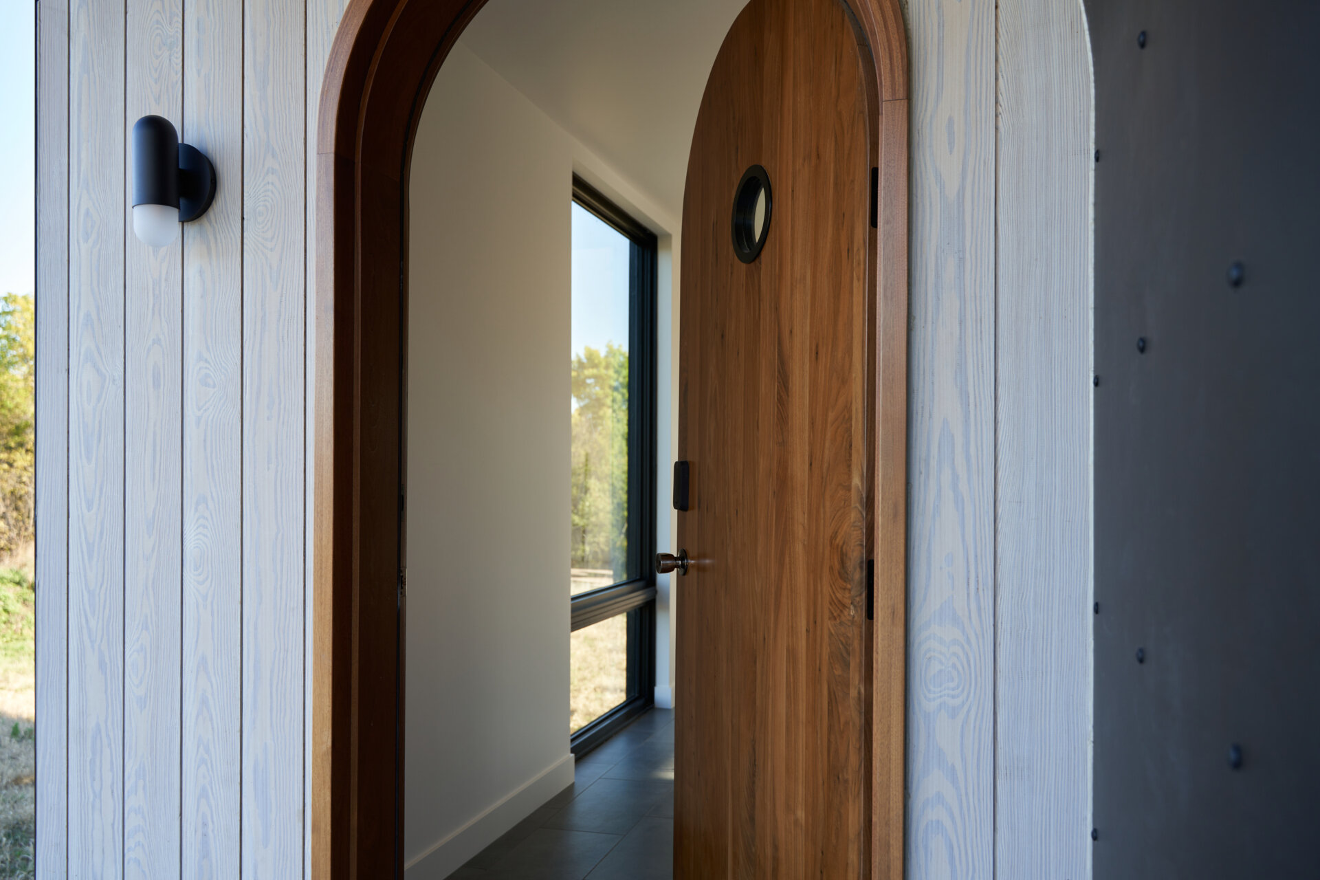 An open arched walnut front door reveals the entrance to a contemporary cabin, with a curved wood frame, pale exterior siding, dark metal porch lining, and a tall window illuminating the hallway.