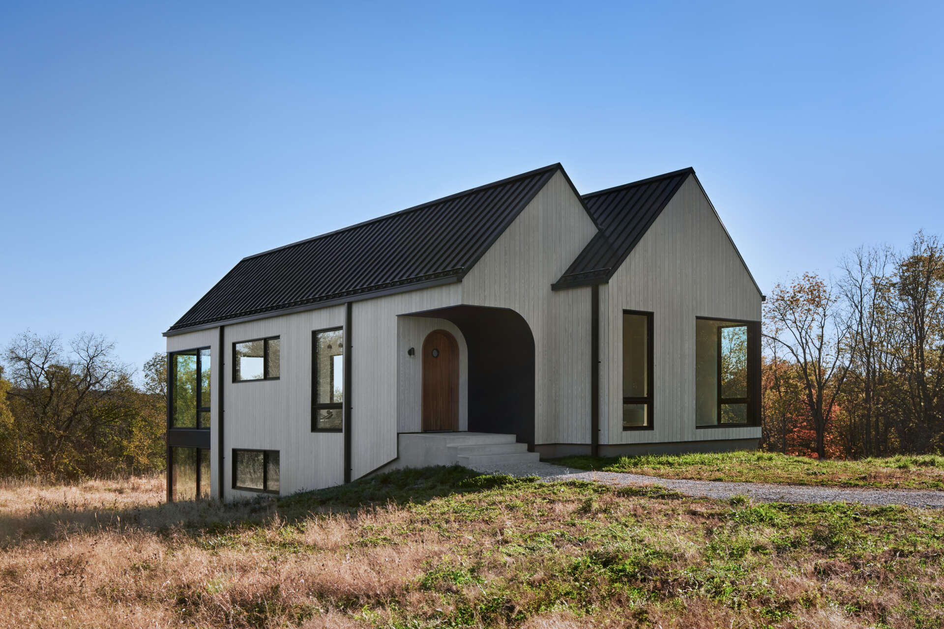 A modern rural cabin with white vertical wood siding, black metal gabled roofs, and a recessed arched front door. The covered entrance sits between two sections of the house, with large windows overlooking the countryside.
