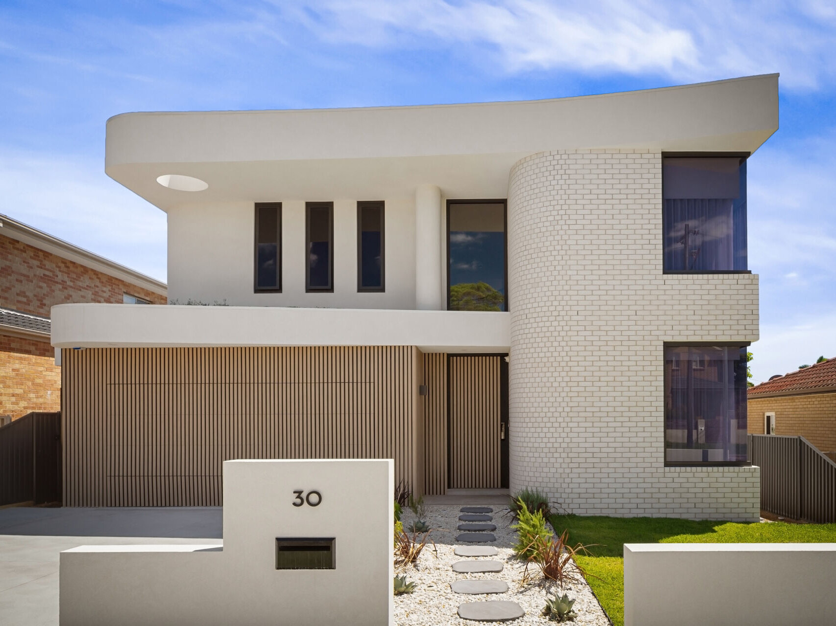 A modern two-story house in Sydney featuring a curved white brick facade, sweeping roofline, and vertical wood battens covering the garage. The contemporary exterior combines rounded architectural forms with tall, narrow windows and a landscaped front entrance.
