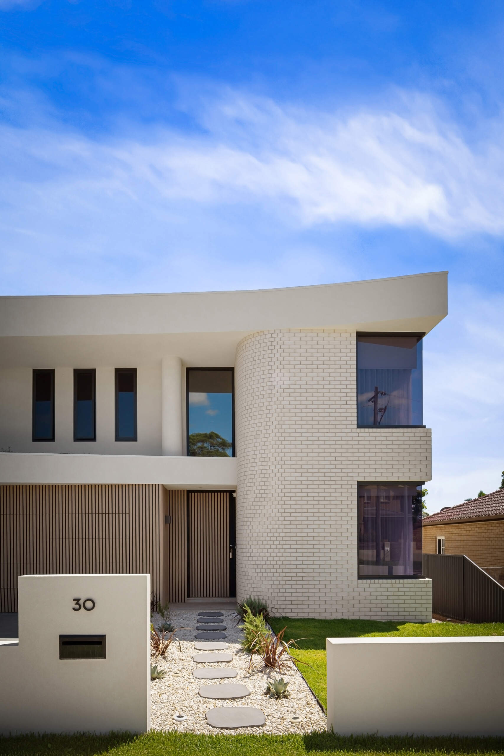 A contemporary Australian house with a two-story curved white brick wall, projecting white roof, and dark-framed windows. A stepping-stone pathway leads through the landscaped front yard to an entrance framed by vertical wood battens.