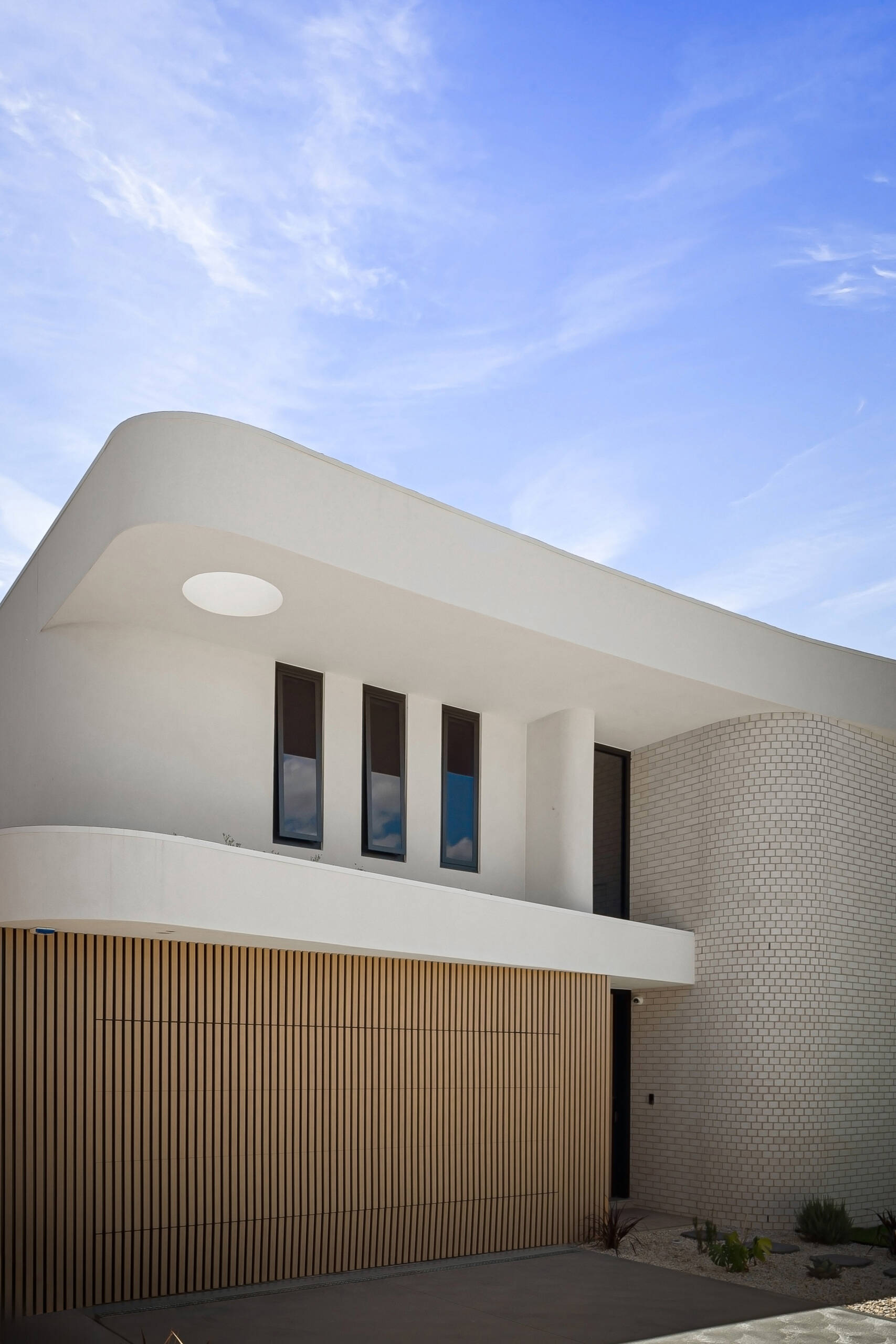 A close-up of a modern house exterior featuring a broad curved roofline, white rendered walls, and a rounded brick facade. Narrow vertical windows, a circular roof opening, and wood battens add architectural detail to the two-story design.