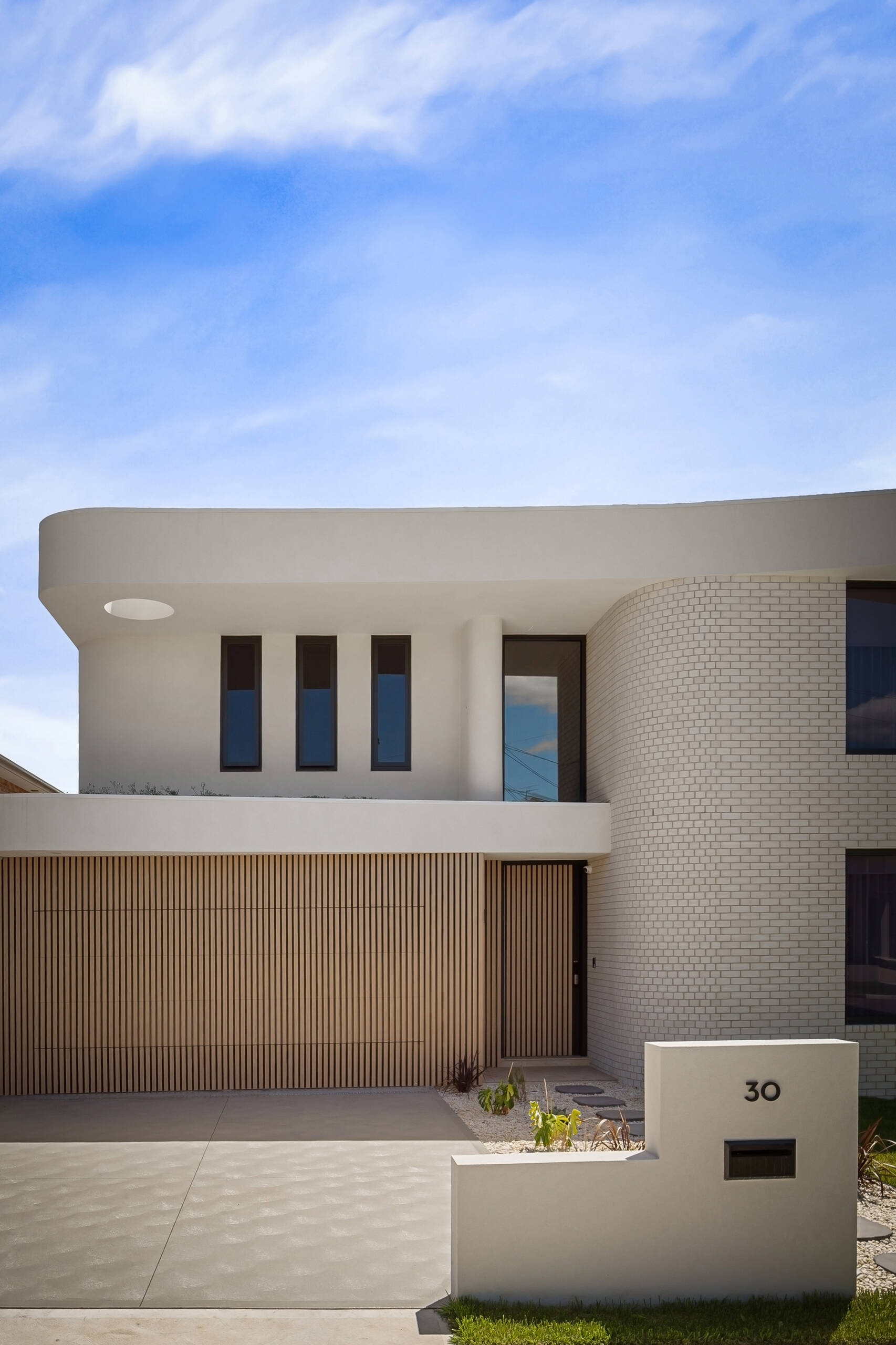 A modern house facade featuring a garage door concealed within a continuous wall of vertical wood battens. A projecting white roof with a circular opening extends above the entrance, alongside a curved white brick wall.