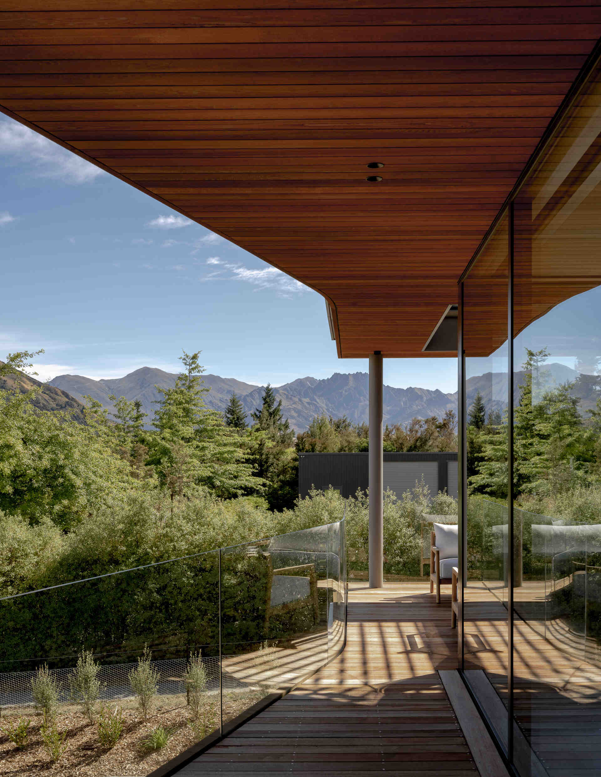 Covered balcony with wood decking, a matching wood ceiling, glass railings, and mountain views. The sheltered outdoor seating area forms part of a contemporary home in W?naka, New Zealand.
