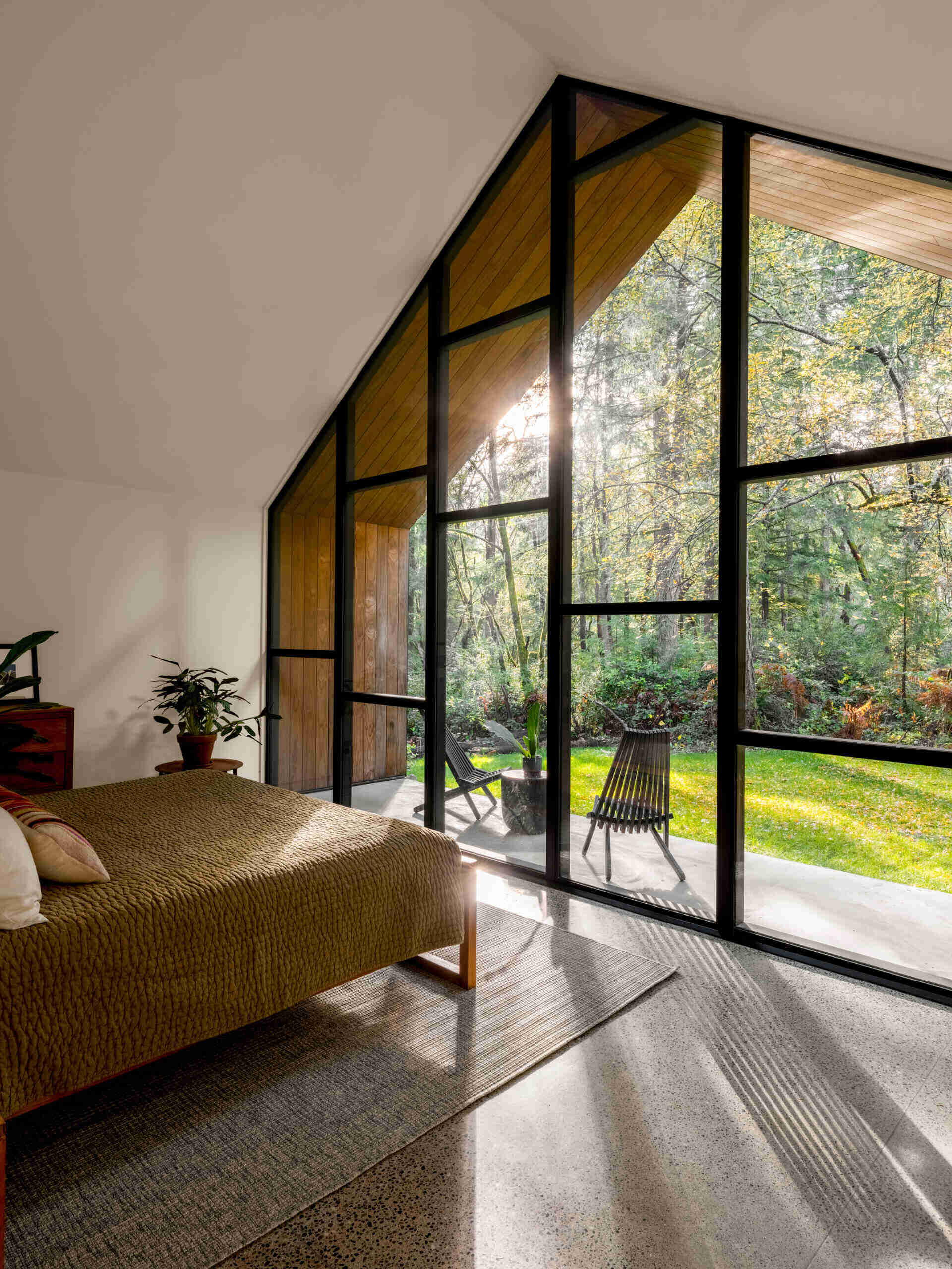 A modern forest bedroom featuring a dramatic gable-shaped glass wall, vaulted white ceiling, low wooden bed, olive-green bedding, and a private outdoor seating area.