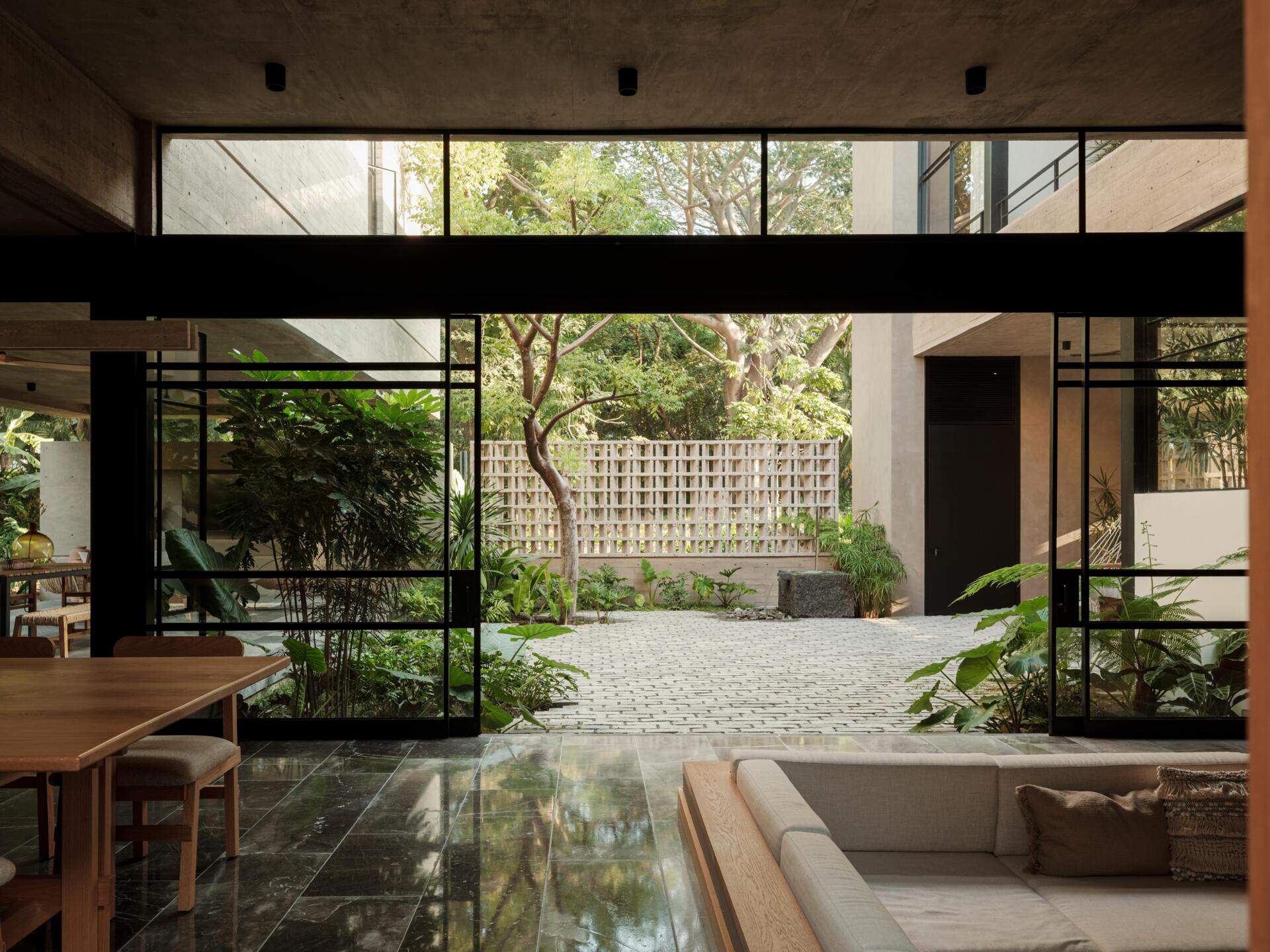 Explore a modern courtyard at Casa Akina in Mexico, featuring mature trees, tropical landscaping, stone paving, a minimalist water feature, geometric breeze-block walls, and a shaded hammock area.