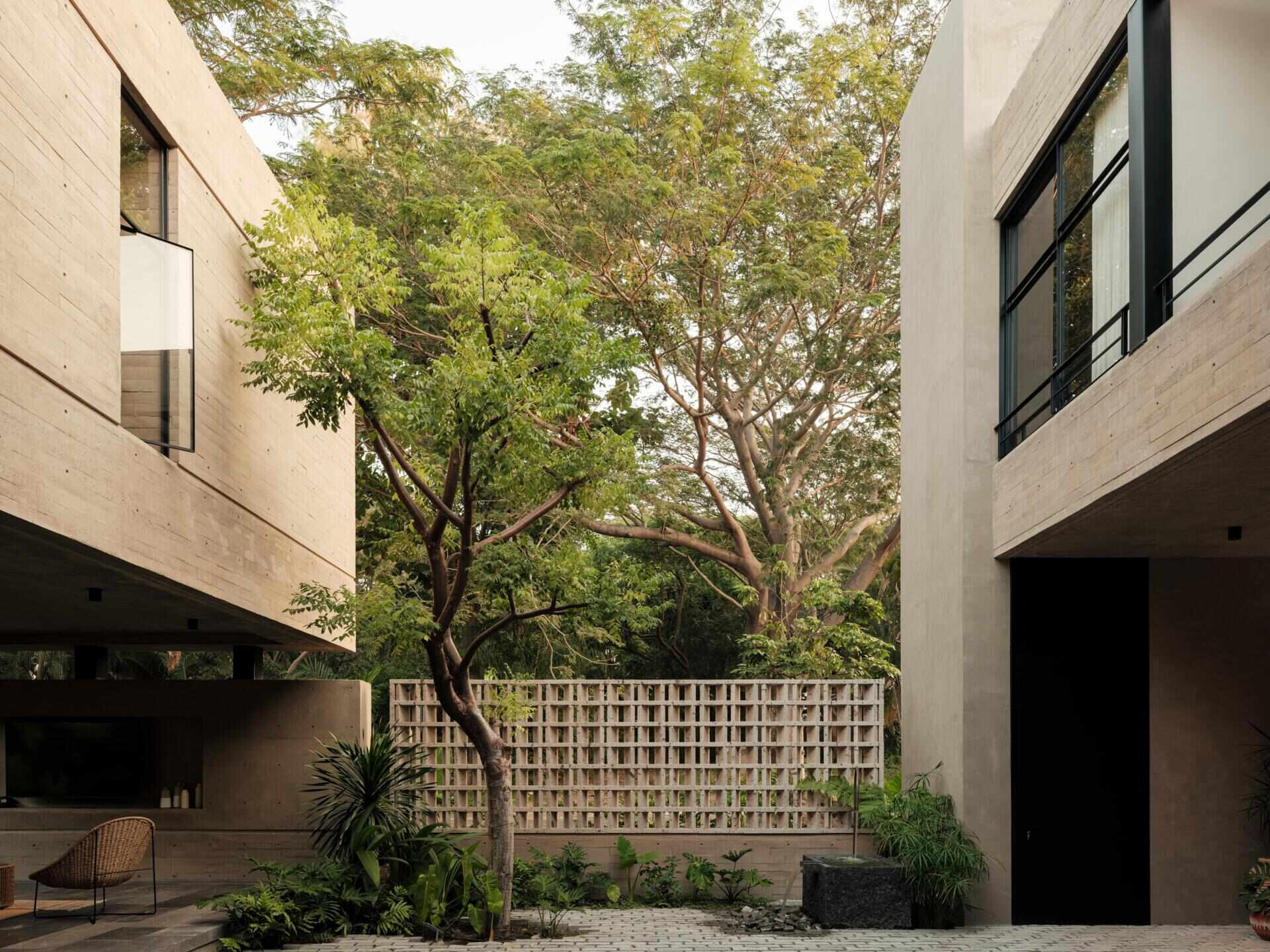 Explore a modern courtyard at Casa Akina in Mexico, featuring mature trees, tropical landscaping, stone paving, a minimalist water feature, geometric breeze-block walls, and a shaded hammock area.