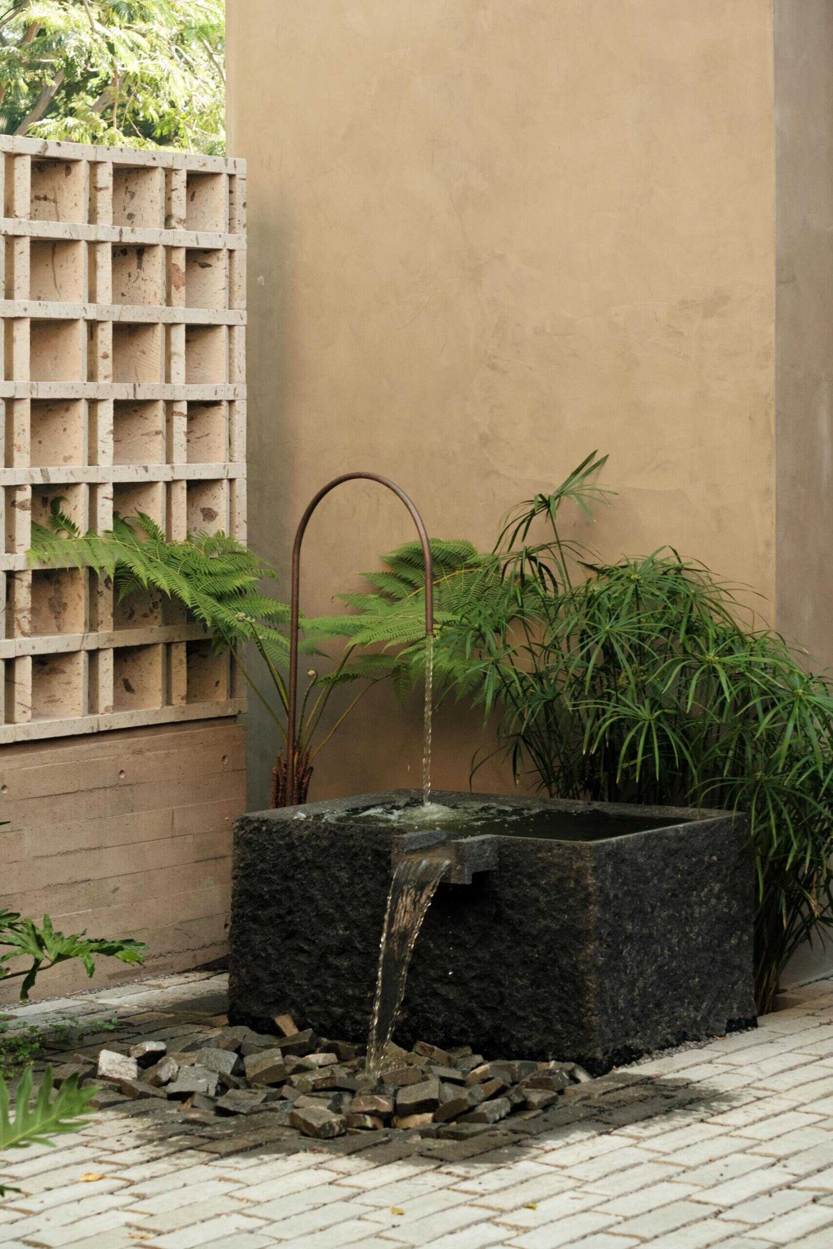 Explore a modern courtyard at Casa Akina in Mexico, featuring mature trees, tropical landscaping, stone paving, a minimalist water feature, geometric breeze-block walls, and a shaded hammock area.