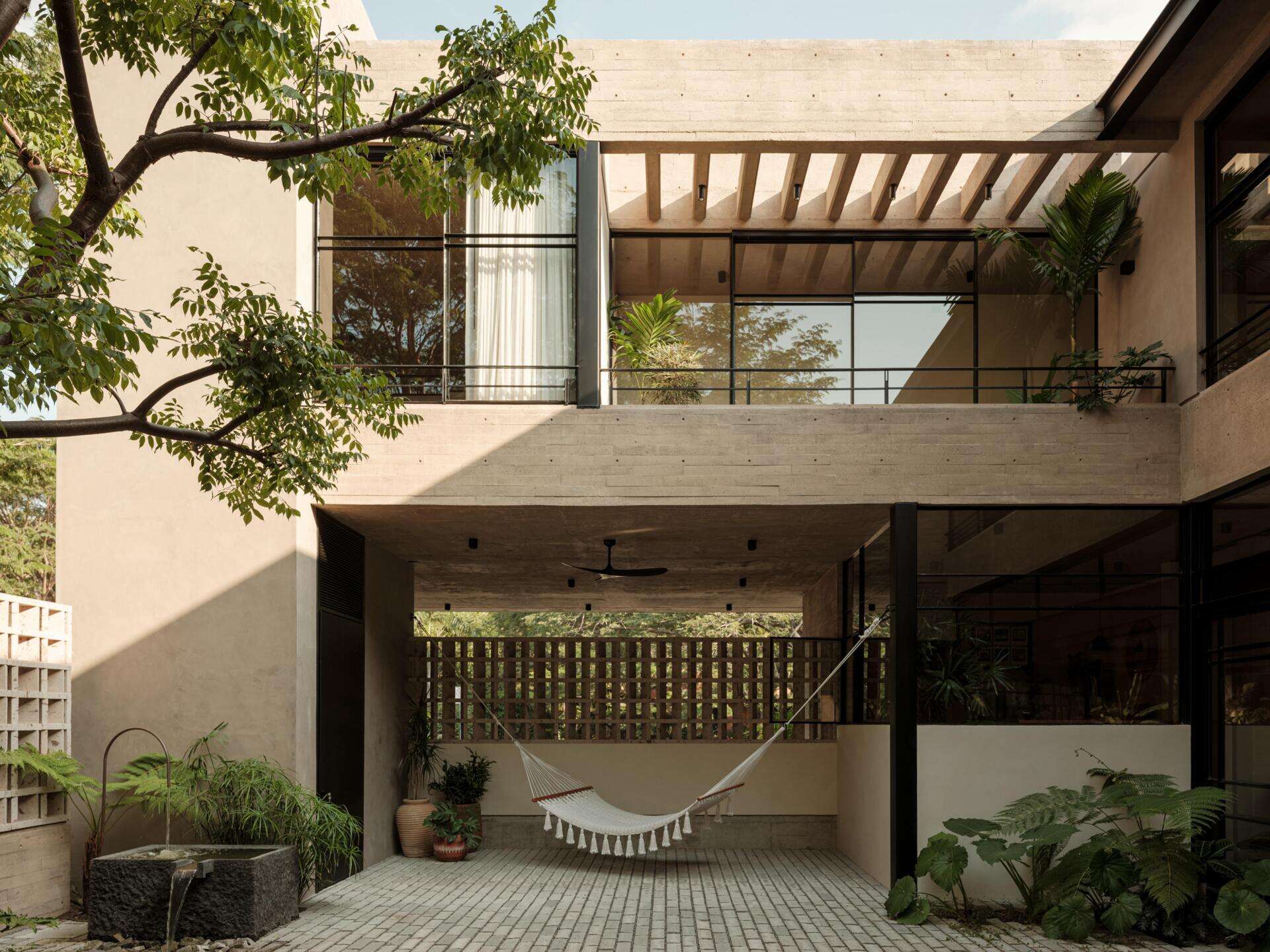 Explore a modern courtyard at Casa Akina in Mexico, featuring mature trees, tropical landscaping, stone paving, a minimalist water feature, geometric breeze-block walls, and a shaded hammock area.