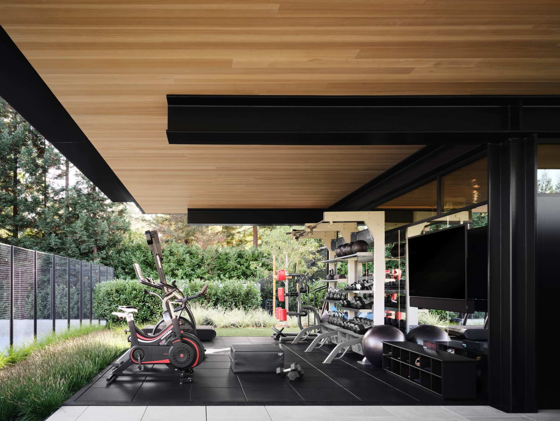 Discover an open-air home gym at a modern California house designed by Olson Kundig. Featuring cardio machines, weight-training equipment, rubber flooring, mirrored walls, and a covered timber ceiling overlooking landscaped gardens.